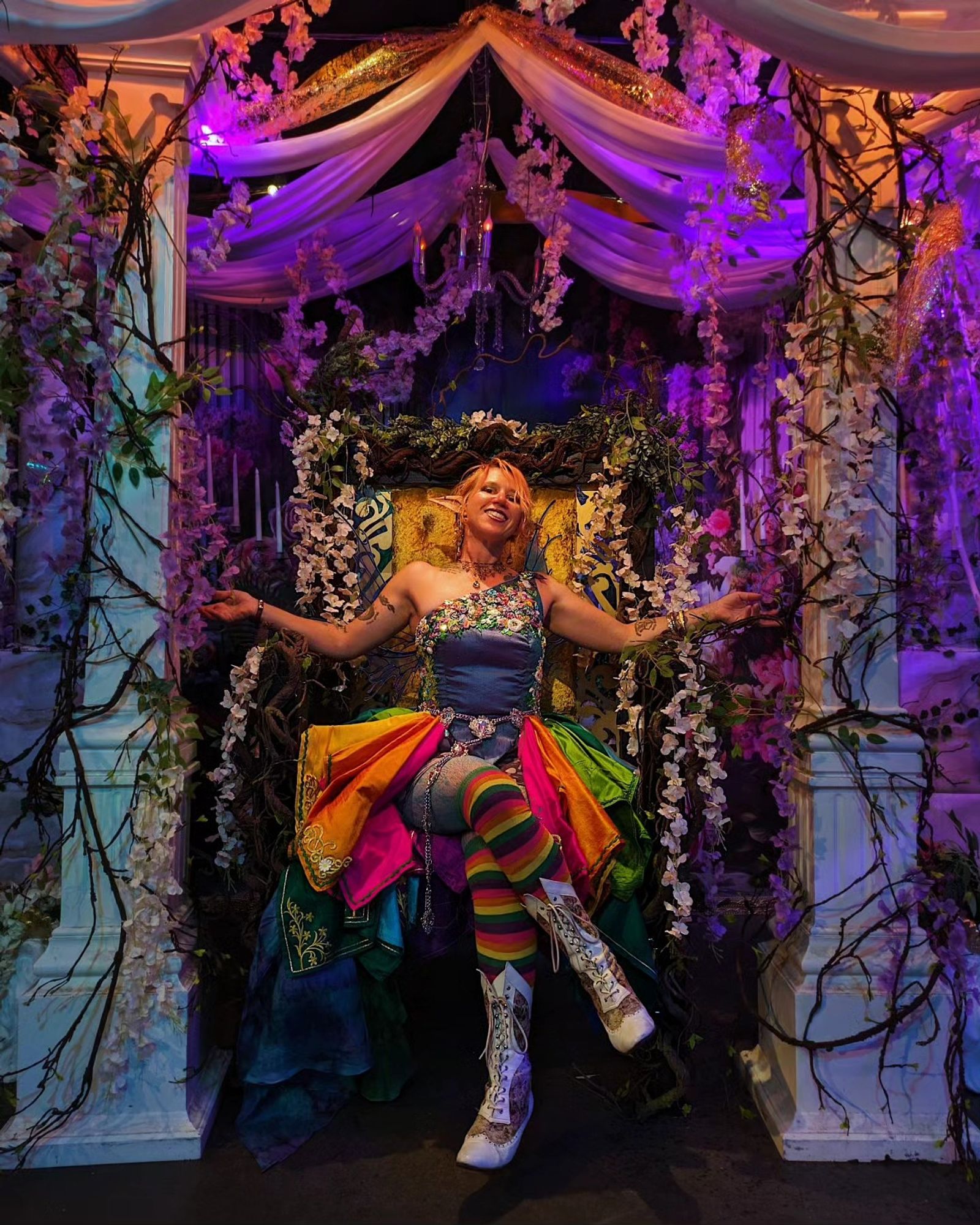 Rowan, Heir of the Seelie Court, born of Spring and Summer, sits on an elaborate throne of flowers and vines, arms splayed out in a regal pose, resplendent in all the colors of the rainbow.  Taken at the Quest of Thrones event by Elevate Immersive.