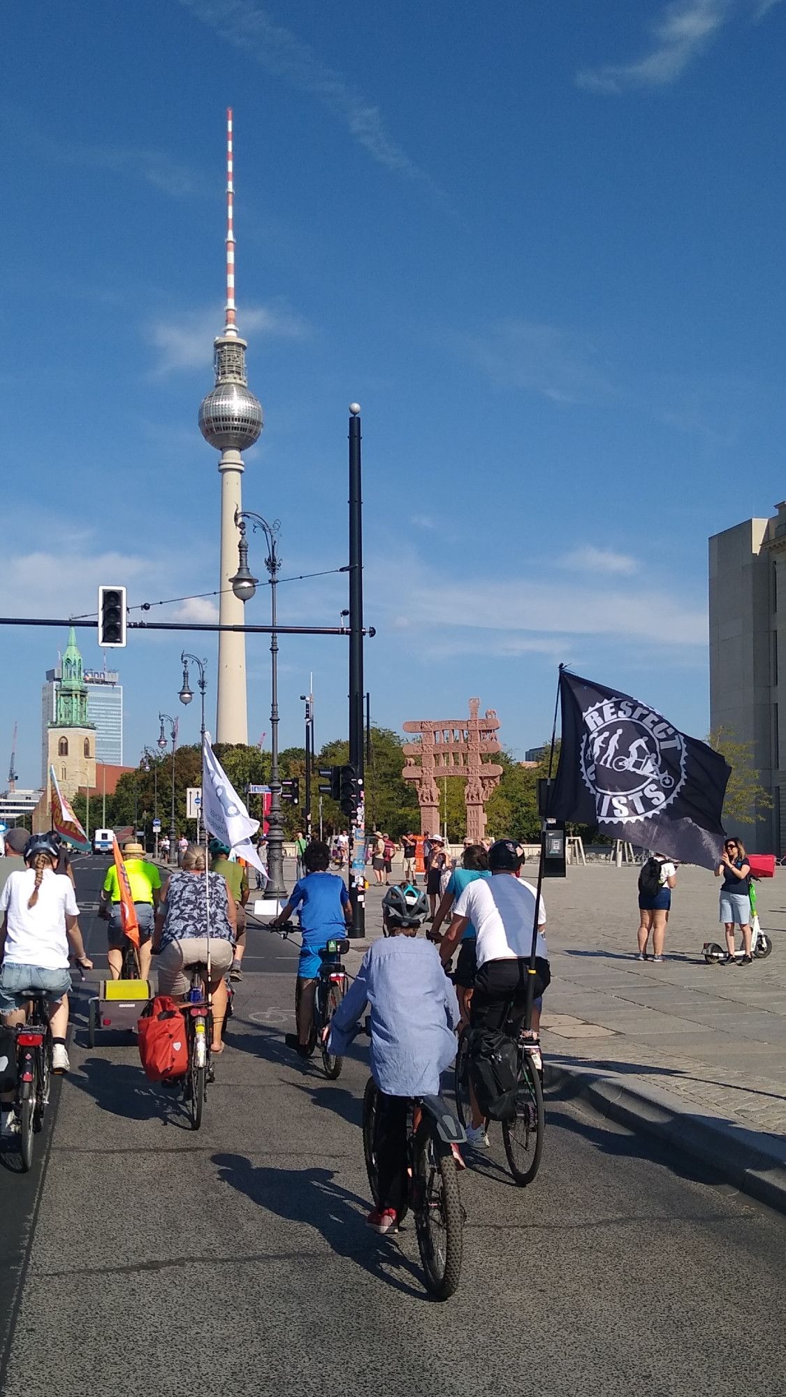 Eine Gruppe Radfahrende fährt am Humboldtforum vorbei Richtung Rotes Rathaus.
Man sieht den Fernsehturm im Hintergrund. Rechts im Vordergrund weht die Respect Cyclists Fahne an einem Fahrrad.