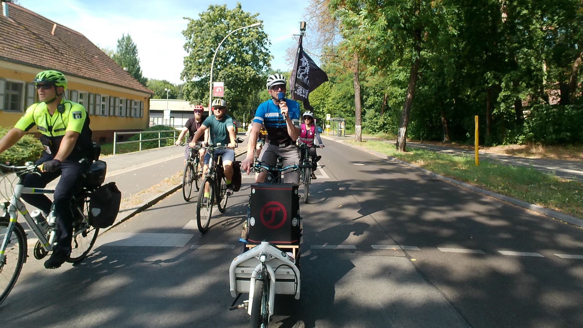 Zentral im Bild, Bosse der auf seinem Lastenrad fahrend die Demo moderiert. Links daneben ein Mitglied der Fahrradstaffel der Polizei Berlin, Im Hintergrund sind weitere Radfahrende zu sehen.