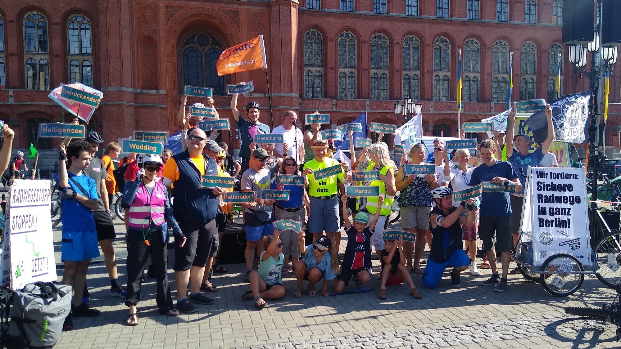 Eine Gruppe von Menschen posiert vor dem Roten Rathaus für die Kamera mit vielen Berliner Ortsschildern in den Händen.
Links im Bild ein Banner mit der Aufschrift "Radwegestopp stoppen!", Rechts im Bild ein Banner: "Wir fordern: Sichere Radwege in ganz Berlin!"
Im Hintergrund sind Fahnen von Cahnging Cities und dem ADFC zu sehen.