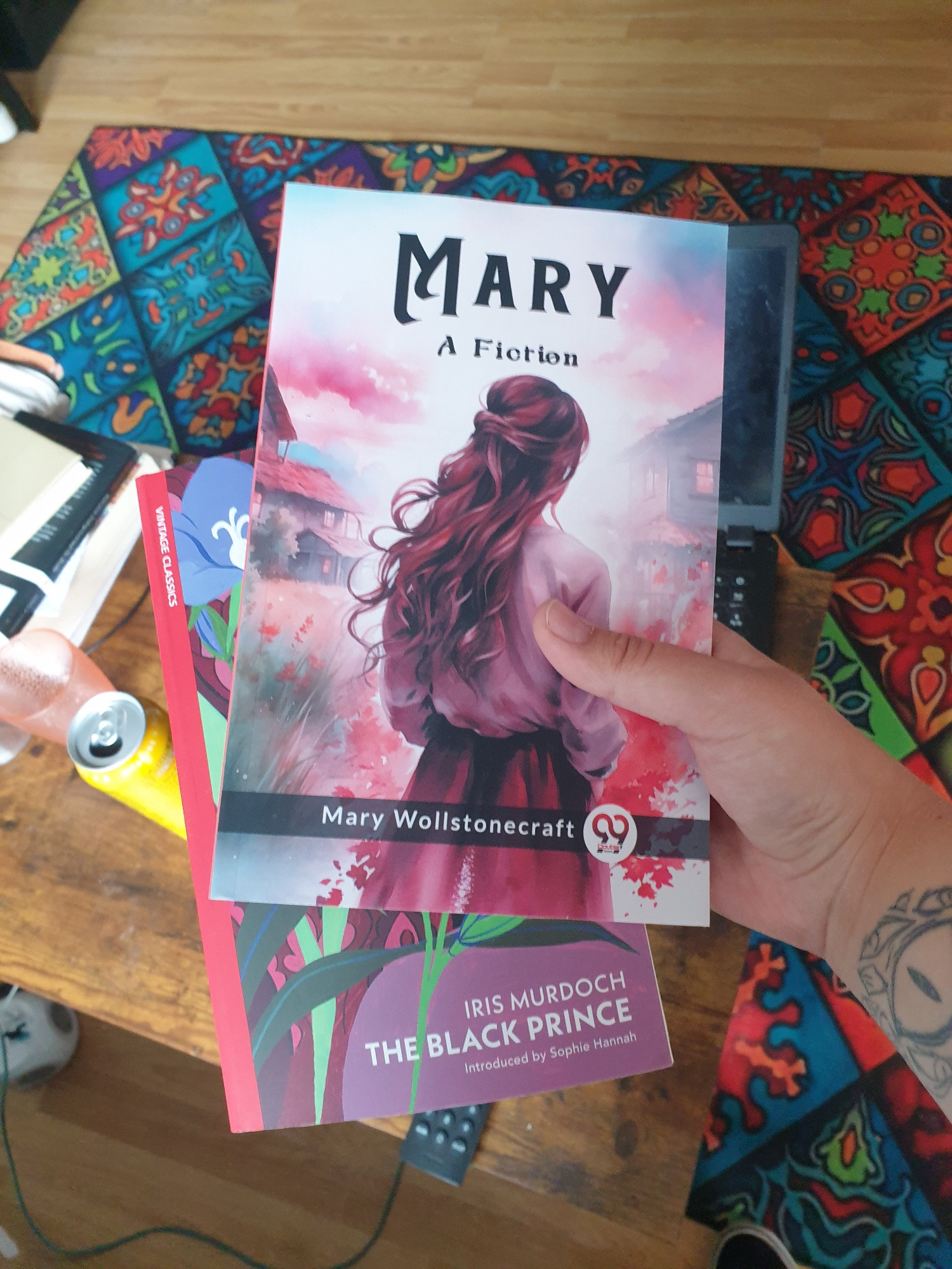 me holding a copy of 'mary, a fiction' by mary wollstonecraft, with a 'double 9 books' logo next to author name (cover shows painting style, back of a woman with long red hair, a white shirt tinted red in the light and a red skirt with houses in the background and a red/purple/blue cloudy evening sky) and beneath it a copy of 'the black prince' by iris murdoch. beneath the title it reads 'introduced by sophie hannah'. Along the binding on the left, there is a red strip which at the top reads 'vintage classics'.