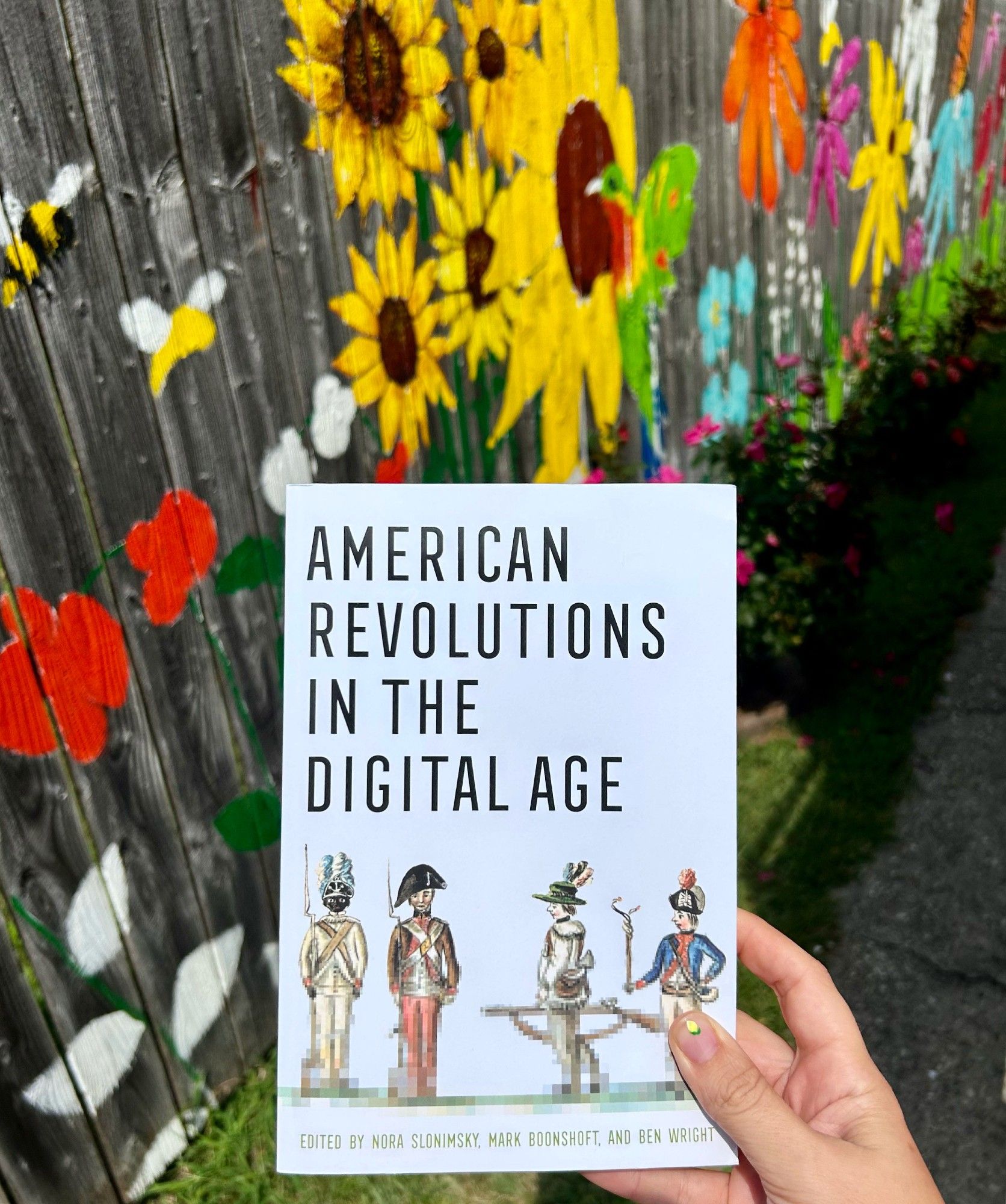 a grey book with a pixelated image of revolutionary soldiers in front of a painted fench with flowers, birds, and bees.