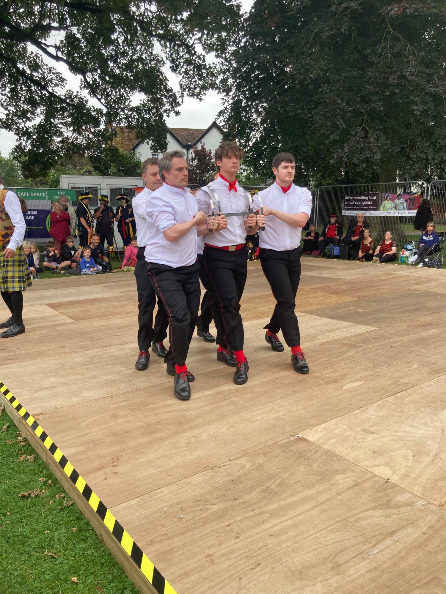 Five men (you can only see four) in white shirts and black trousers with red trimming, dancing together in rapper dance, a folk dance from North East England, on a wooden stage outdoors with people watching