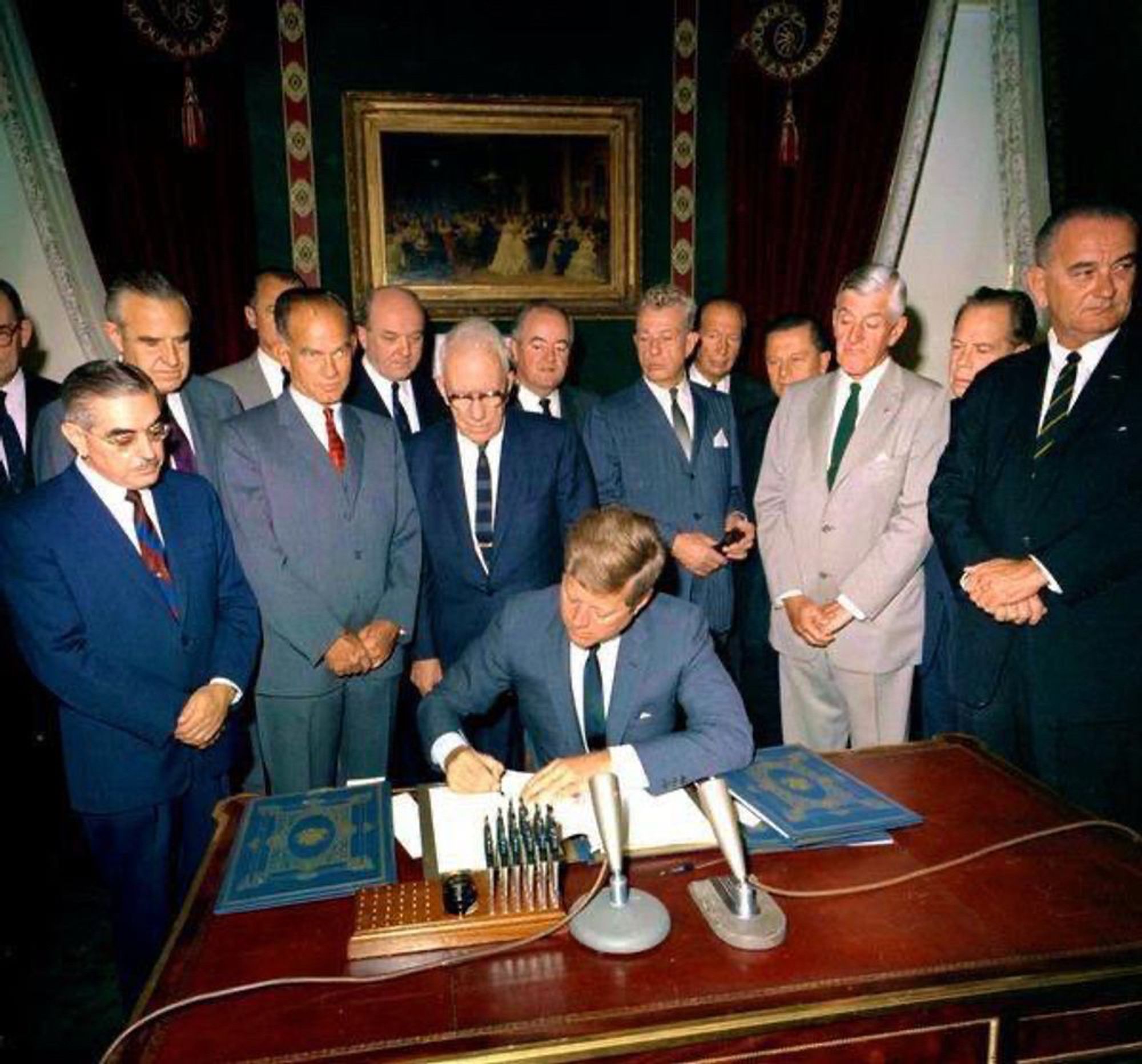 A color photograph of President John F. Kennedy sitting at a desk in the White House and signing a copy of the Limited Test Ban Treaty, as a group of senators stands behind him. Vice President Lyndon Johnson stands at the far right in the dark suit, hands clasped.