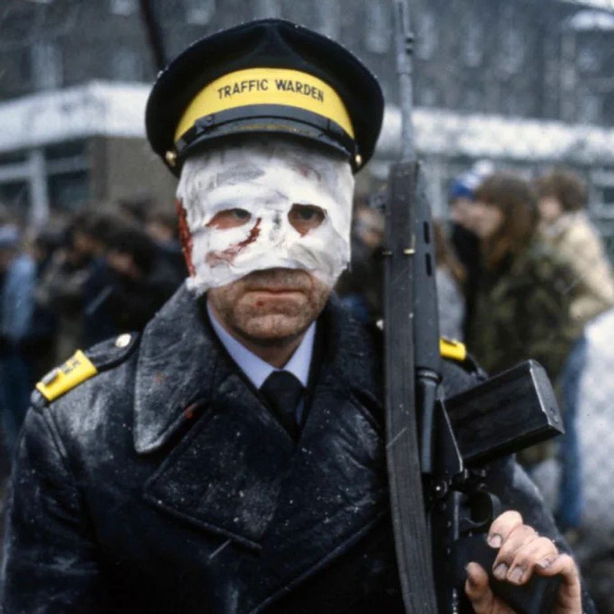 A publicity still from "Threads" showing traffic warden, his upper face heavily bandaged and bloodied, his uniform dirty, holding a semiautomatic rifle against his left shoulder.