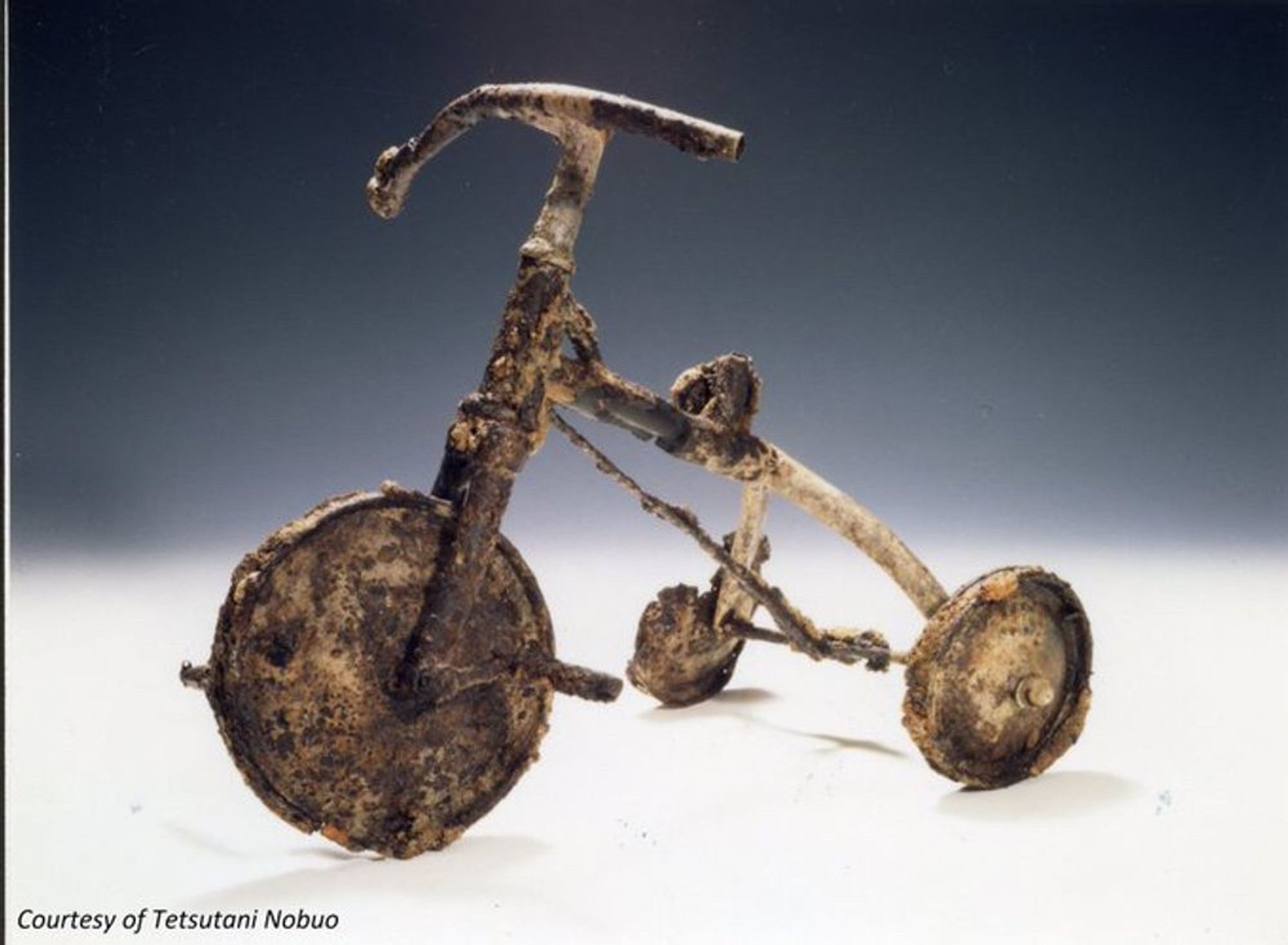 A color photograph of a damaged and partially melted children’s tricycle in the Hiroshima Peace Memorial Museum. On the morning of August 6, 1945, Shinichi Tetsutani, who was one month shy of turning four years old, was riding this tricycle, playing with his best friend, a girl named Kimi, 1,500 meters away from where the Little Boy atomic bomb exploded overhead. When found in the wrecked of his house, he was still grasping the red handlebar grip from his tricycle. Severely injured and burned all over his body, calling out, “Water, I want water,” he died that night. Shinichi’s father buried him in the backyard with his favorite toy, and his best friend Kimi, so he could ride it and play even after death. Forty years later, in 1985, Shinichi’s father, Nobuo, decided to rebury his son in the family’s gravesite. Unearthing the backyard grave with Kimi’s mother, Nobuo rediscovered the tricycle and donated it to the museum.