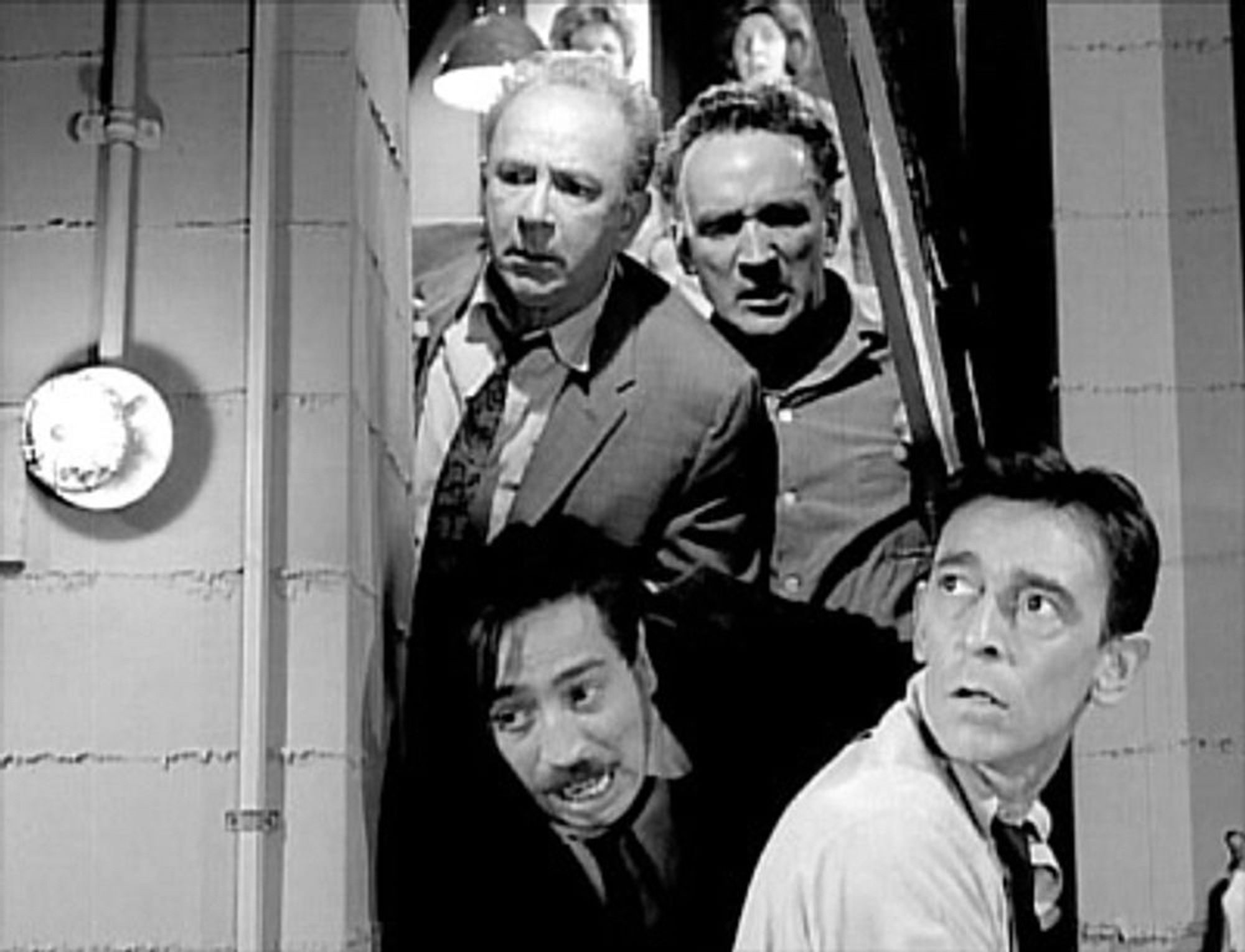 A black and white screenshot of a scene from “The Shelter” showing four men looking through the now broken open door of the shelter, fear and uncertainty on their faces. In the background, two of their wives can be seen with similar expressions on their faces.