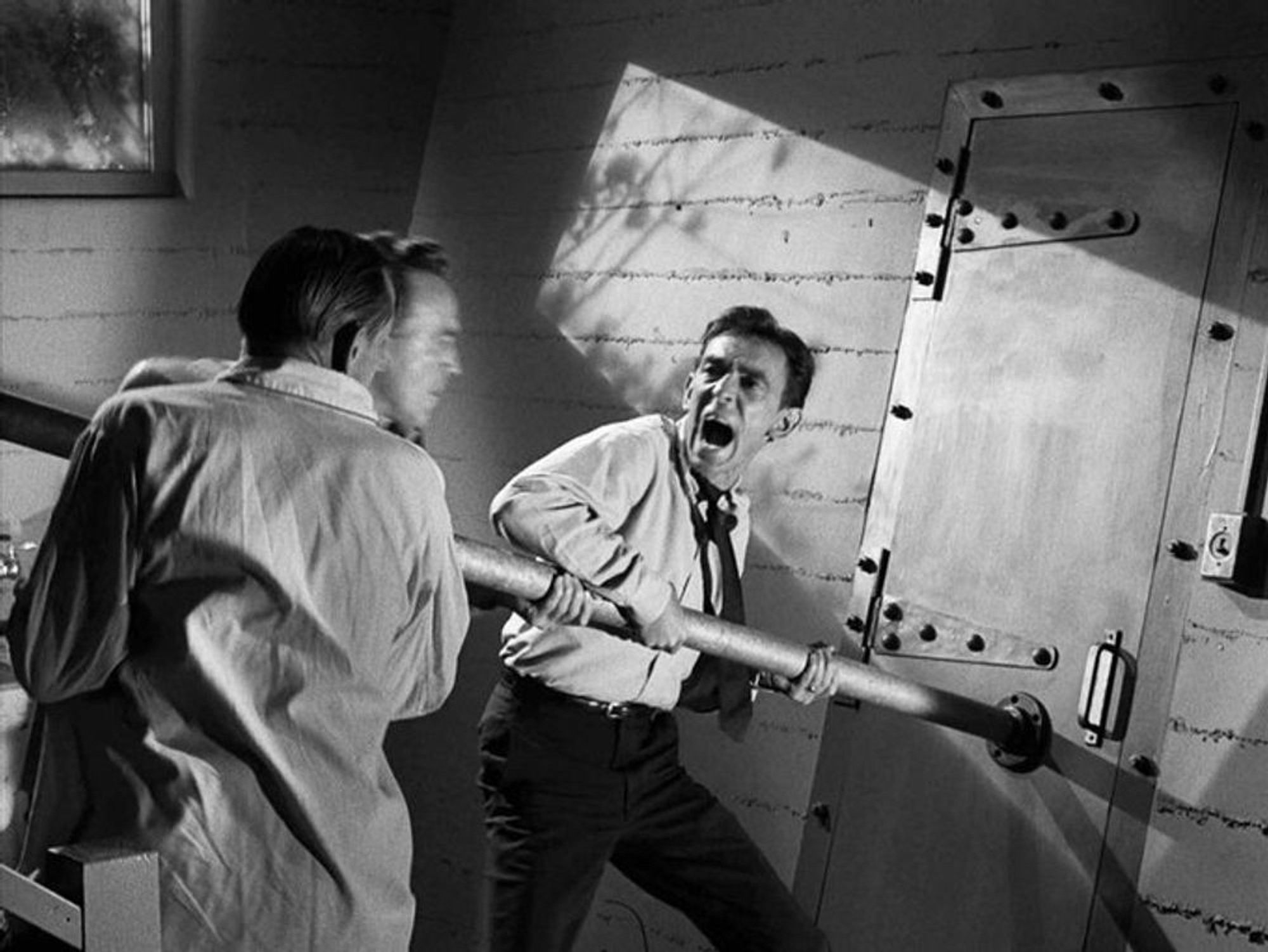 A black and white screenshot of a scene from “The Shelter” showing three men frantically trying to use a large pipe to ram through the reinforced metal door of the shelter.