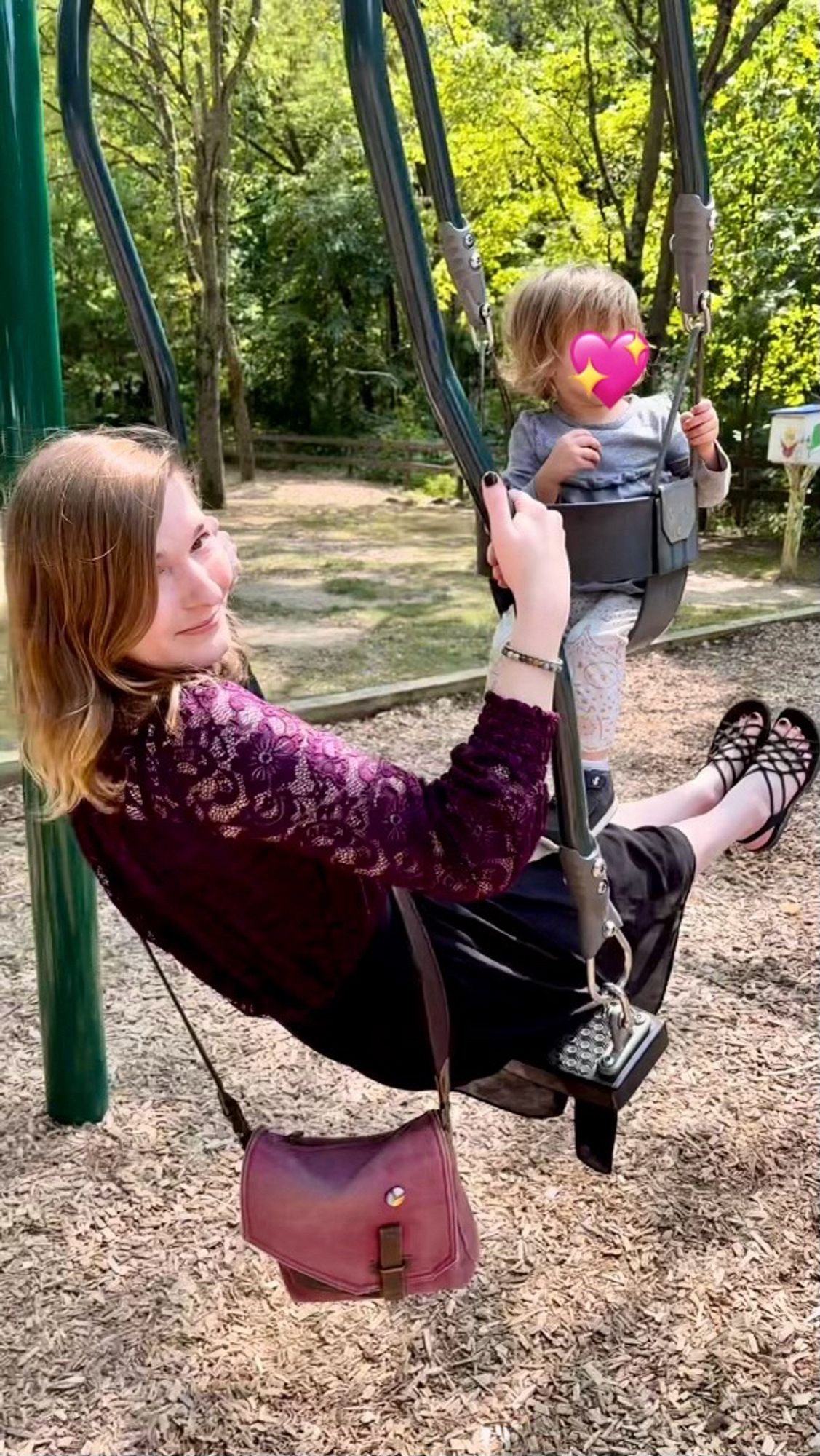 A picture of me on a tandem swing set at a park. I am wearing a purple shirt with mesh floral sleeves and a black skirt with crisscross black string sandels and my faded maroon canvas purse. My child is in a seat attached to my swing, their face is covered with a heart emoji.