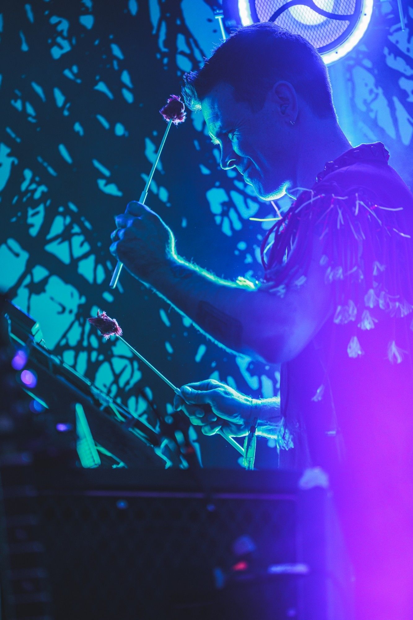 A photo of a male performer playing a percussion instrument in profile lit by blue and purple concert lighting.