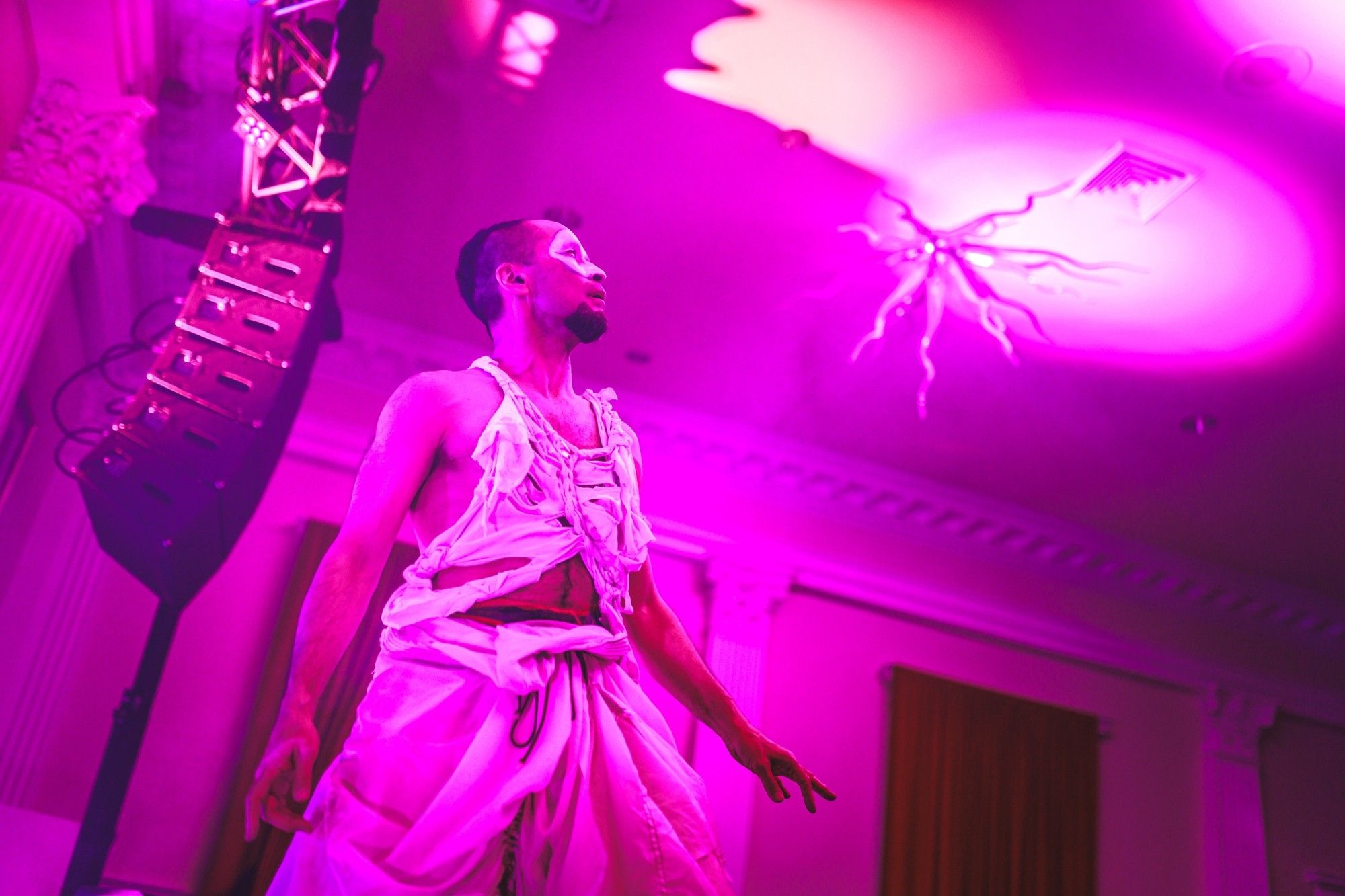 A photo of a male performer in face paint taken from below, posed with his arms held out at his sides looking up at a ballroom ceiling, lit by pink concert lighting.