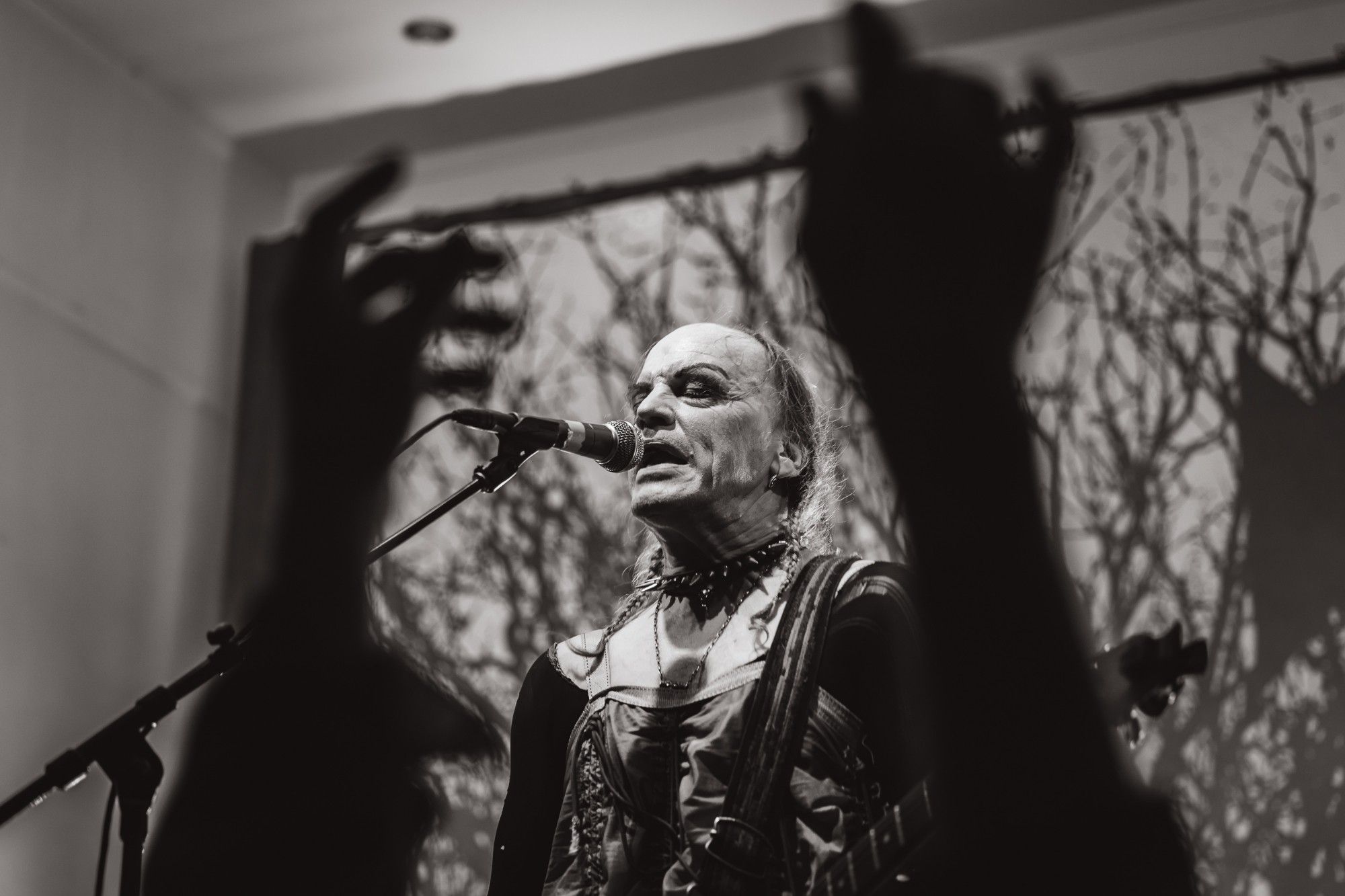 A black and white photo of a male performer in face paint singing into a microphone, framed by a pair of hands in the foreground that are out of focus and in shadow.