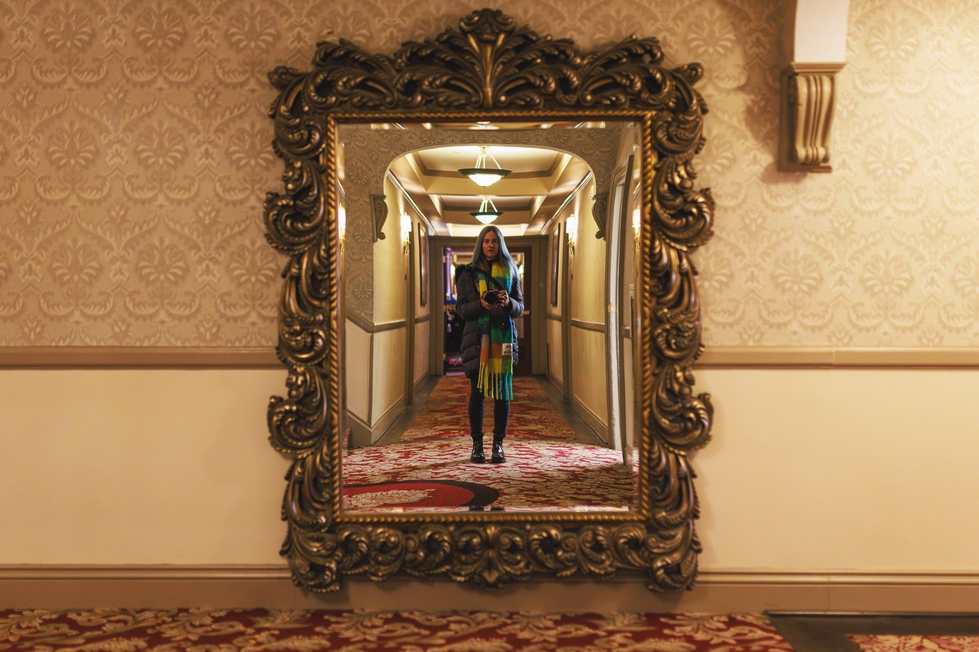 A photo of a woman wearing a coat and scarf taking a selfie in a large ornate mirror.