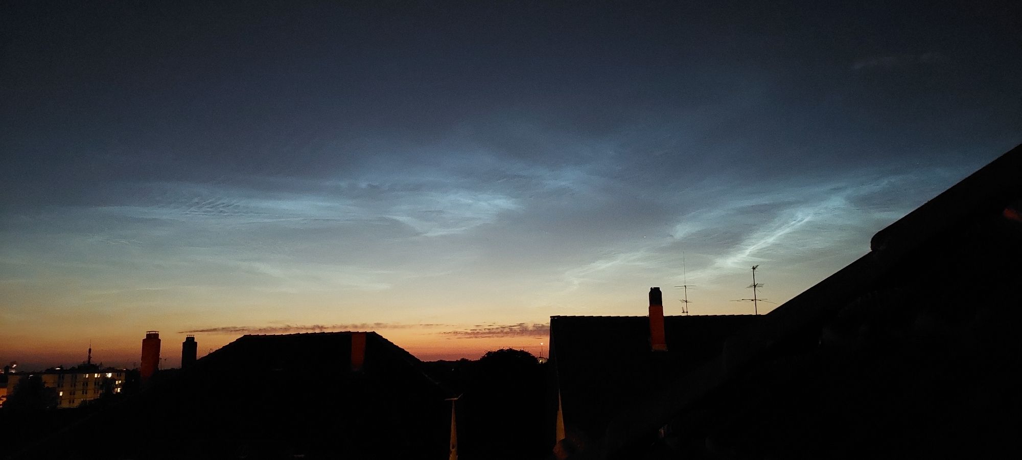 Eisblau leuchtende fein faserige Nachwolken, die sich am dunklen Nordhimmel über dem noch immer vom Abendrot hellen Horizont abzeichnen. Im Vordergrund pechschwarz Dächer der Vorstadt.