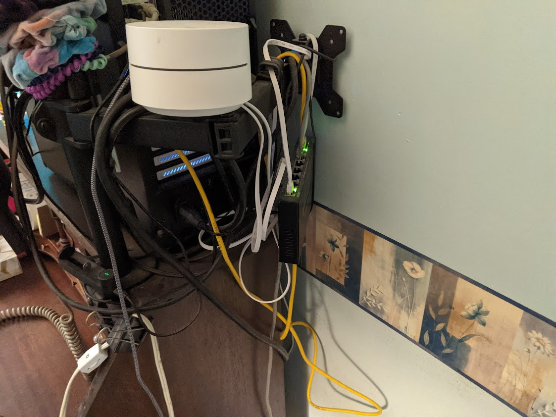 photo of a mess of wires and an ethernet switch hanging from a monitor arm with a google wifi point balancing on it, behind a computer tower perched on the top shelf of a desk