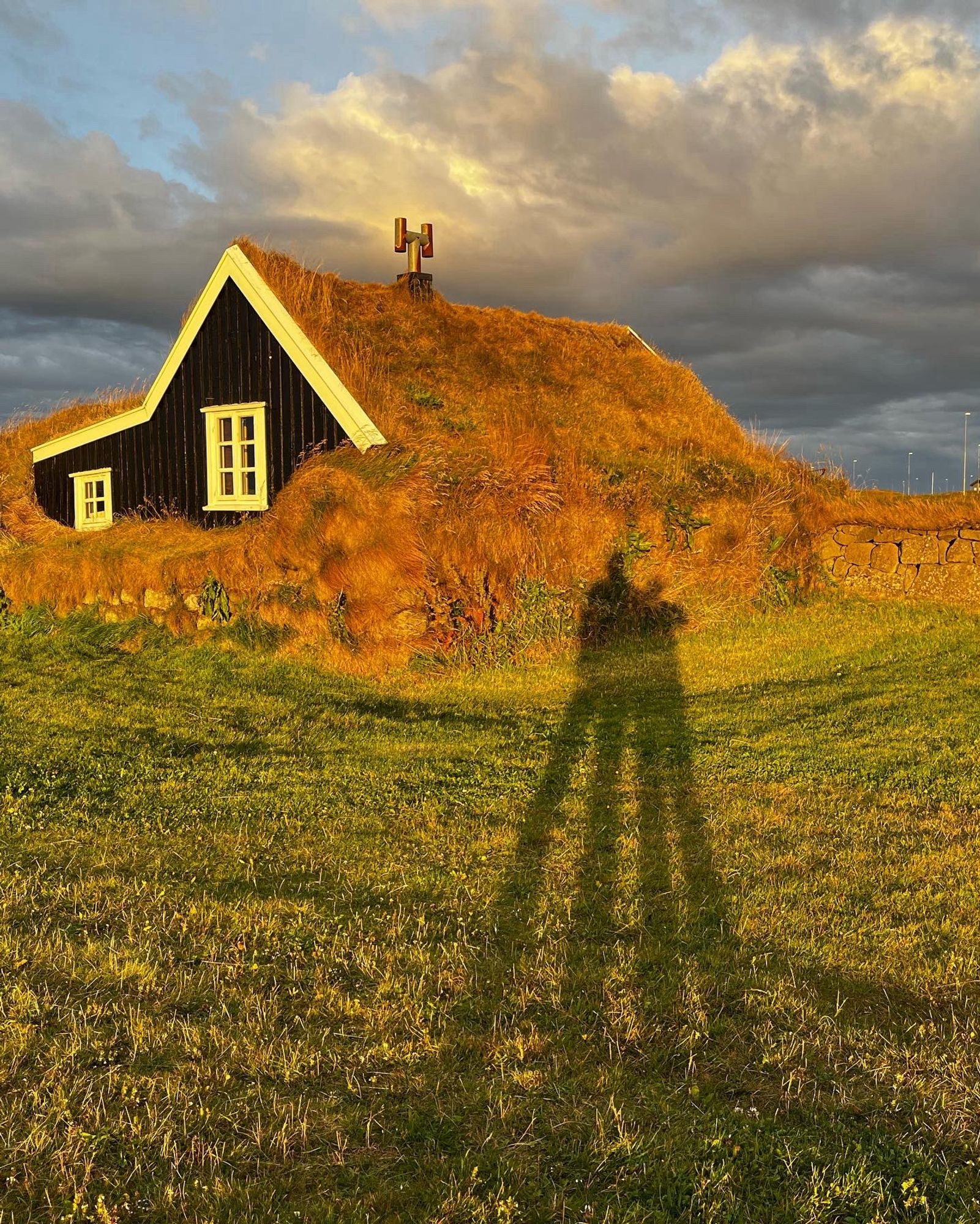 Das Bild zeigt ein traditionelles Torfhaus mit einem mit Gras und Vegetation bedeckten Dach und Seiten. Das Haus hat eine schwarze Außenwand mit weiß gerahmten Fenstern und ist teilweise in einen grasbewachsenen Hang eingebettet. Auf der Spitze des Hügels befindet sich eine einfache Kreuzstruktur, die auf eine historische oder kulturelle Bedeutung des Ortes hinweisen könnte. Der Himmel ist teilweise bewölkt, und das Sonnenlicht wirft den langen Schatten einer Person, die das Foto macht, auf das grüne Gras im Vordergrund, was darauf hindeutet, dass die Sonne tief steht, möglicherweise am frühen Morgen oder am späten Nachmittag. Das Licht verleiht der Szene einen warmen, goldenen Farbton und betont besonders die Texturen des Grases und der Hausseite.