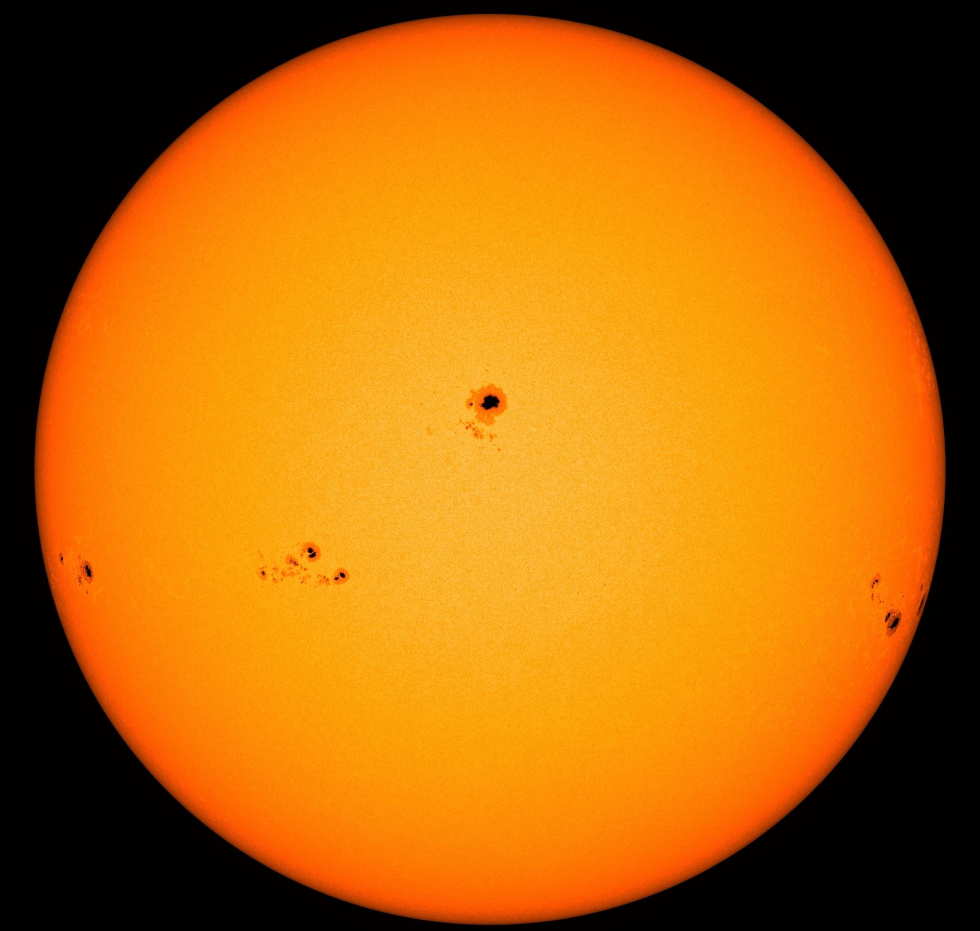 Die erdzugewandte Seite der Sonne in gelber Farbe, die in Richtung des Randes ins Rötliche übergeht. Genau in der Mitte ein größerer dunkler Sonnenfleck und auch darunter mehrere deutlich kleinere Fleckengruppen.