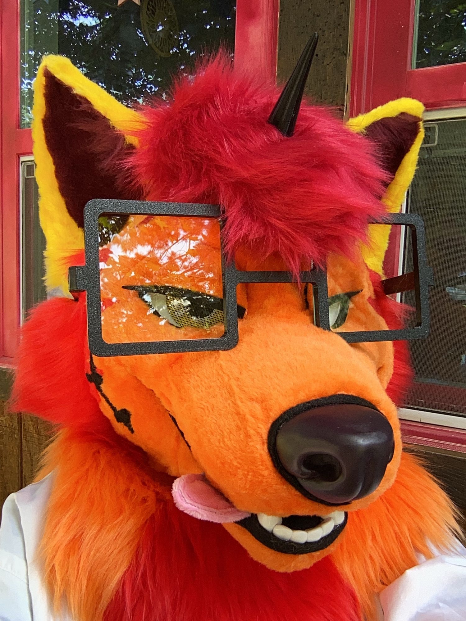 Photo of an orange, red and yellow Tibetan fox fursuiter with a unicorn horn and large glasses. They have magnetic eyelids, and their tongue sticks out to the side.