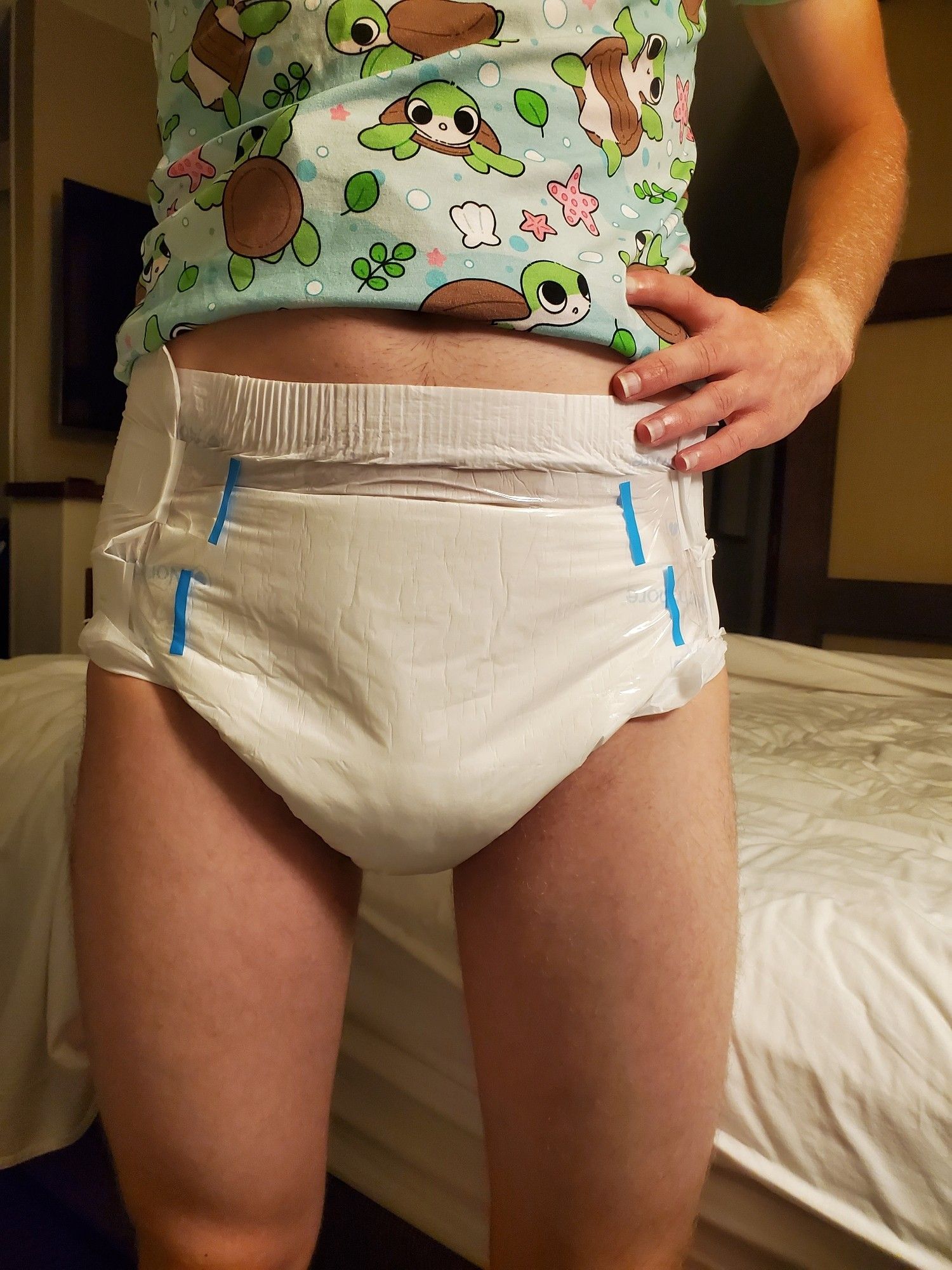 Boy standing next to his bed wearing cute baby turtle printed onesie that he's turned up so you can clearly see his clean white diaper front.