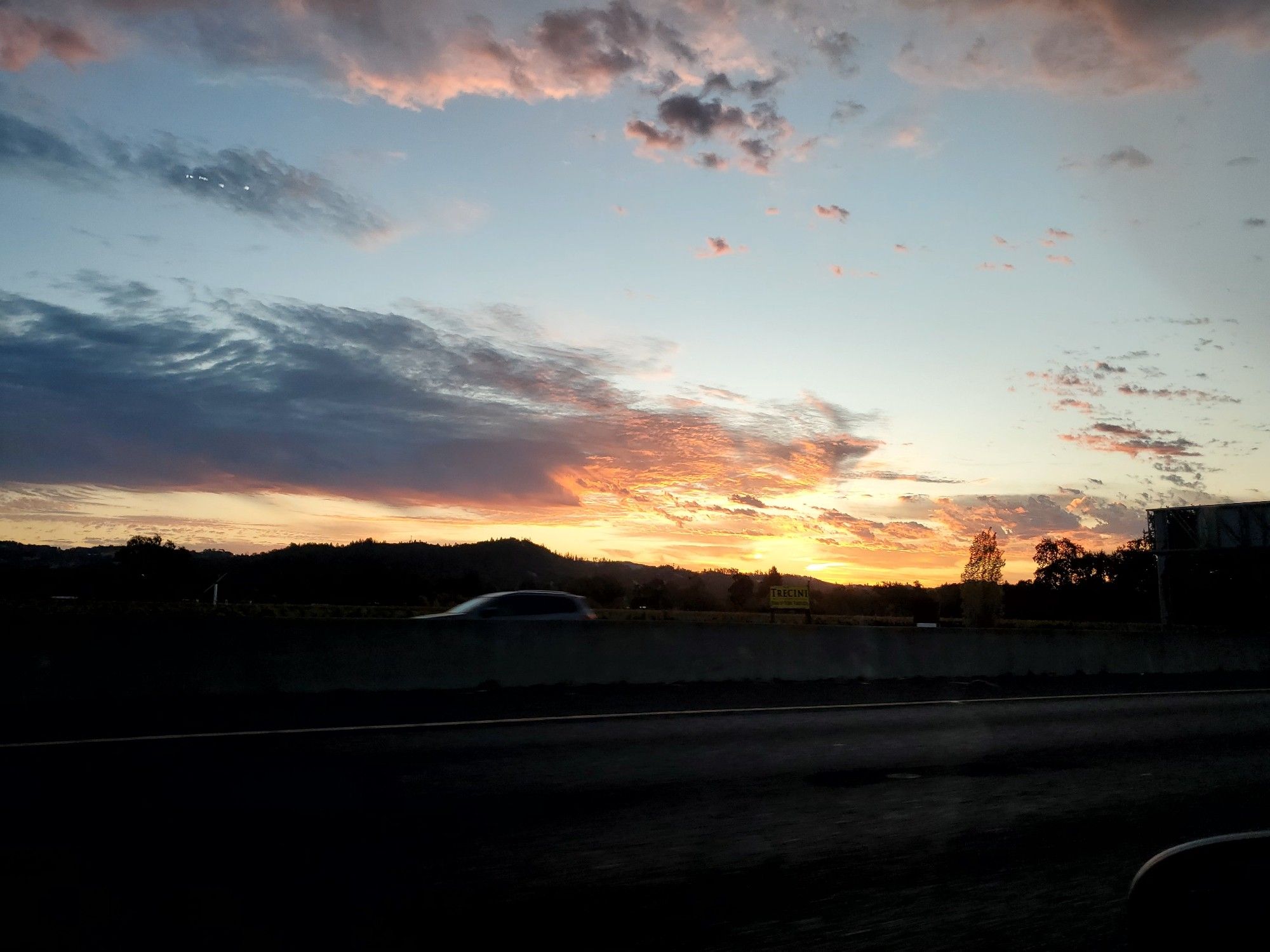 Orangish yellow sunrise bouncing off the clouds as the sun comes over the hill, revealing a pale blue sky above a still darkened landscape.