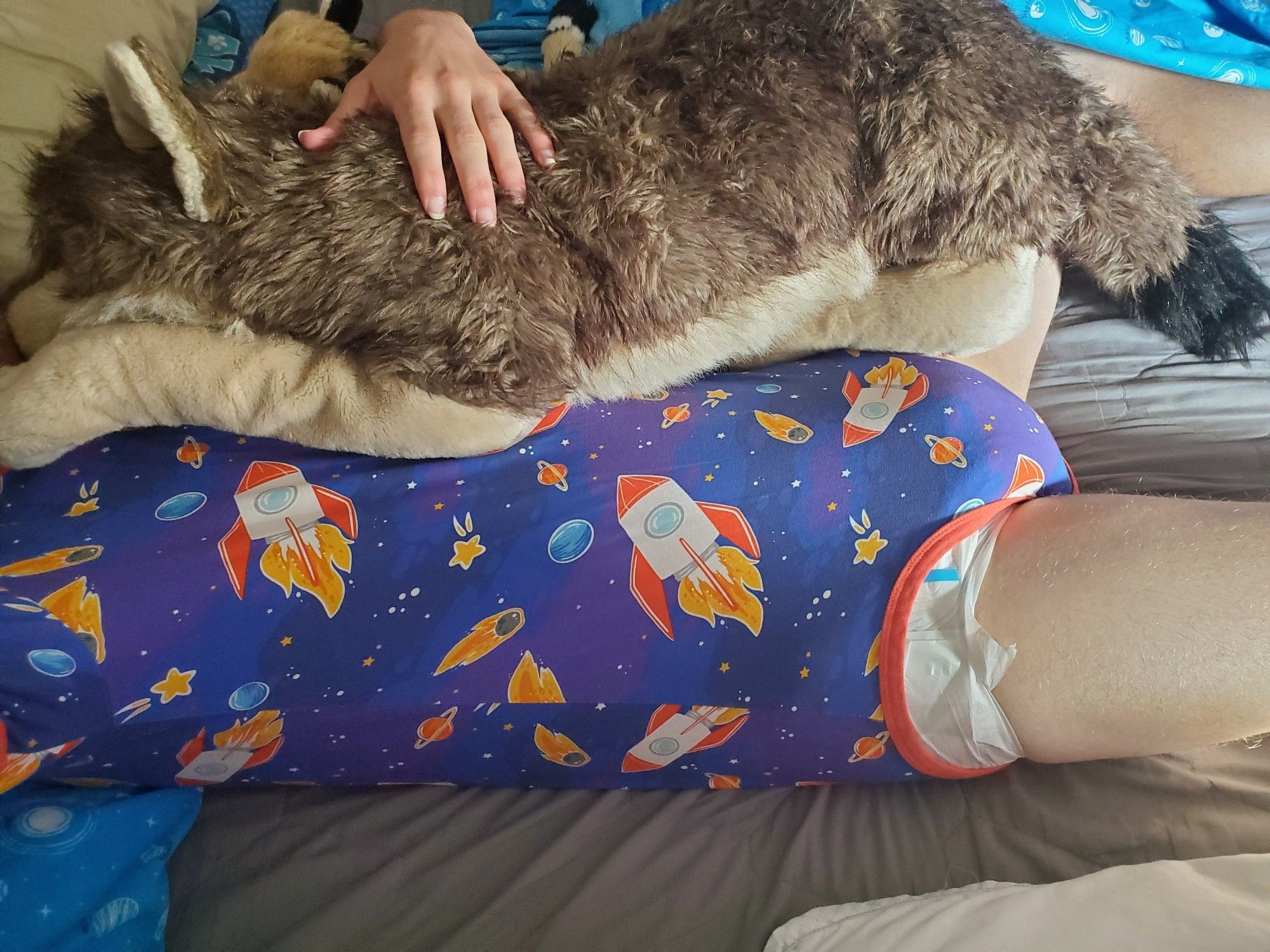 A sleepy boy laying in bed, wearing purple and blue space and rocket ship themed onesie secured snuggly around is plain white diaper, holding onto his wolf plush that's as big as he is, sunlight beaming in through the window. #abdl #irldiaper #babyfur