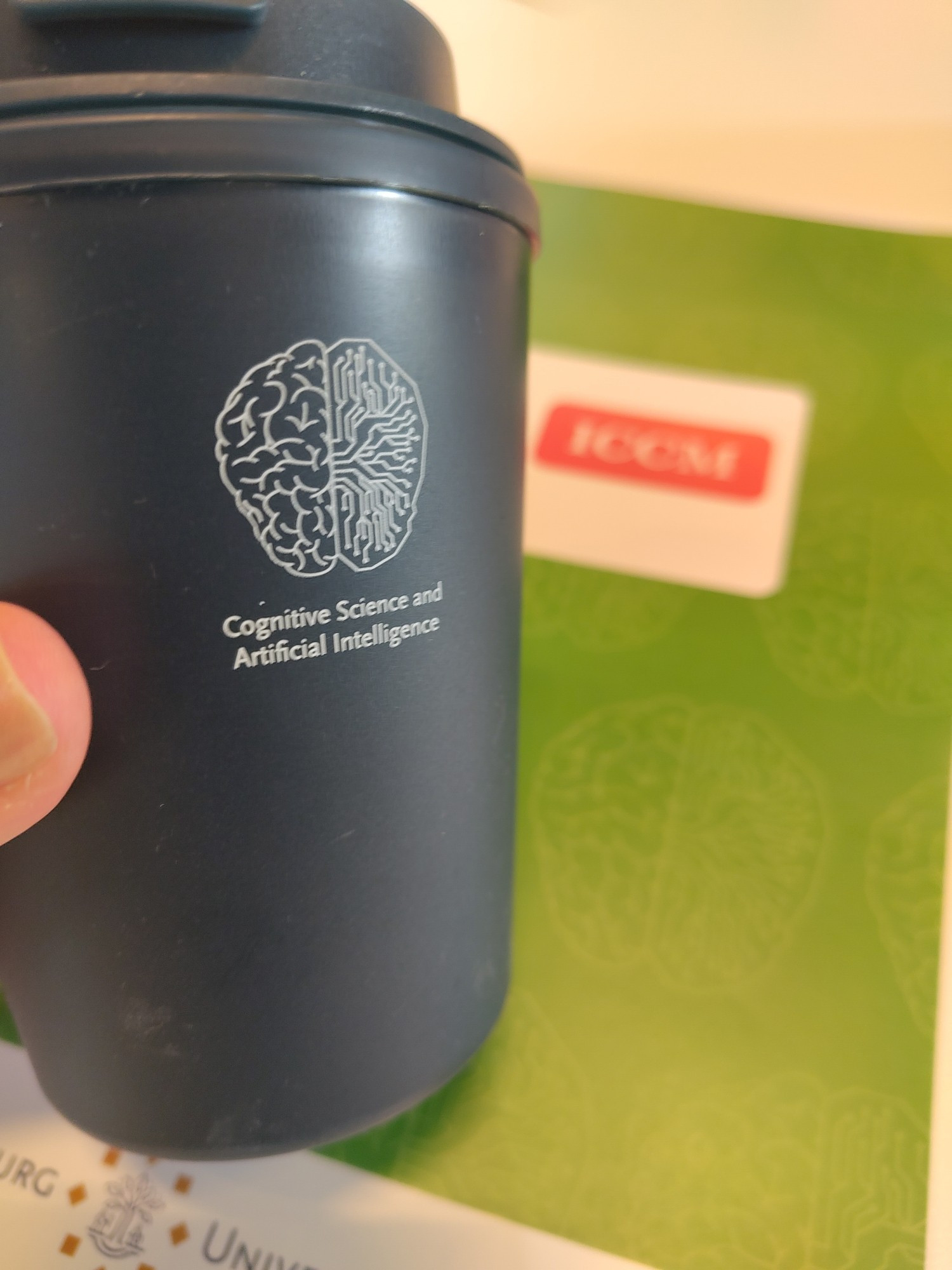 A reusable coffee mug with a printed on brain, which is half stylized circuits, held in front of a conference program