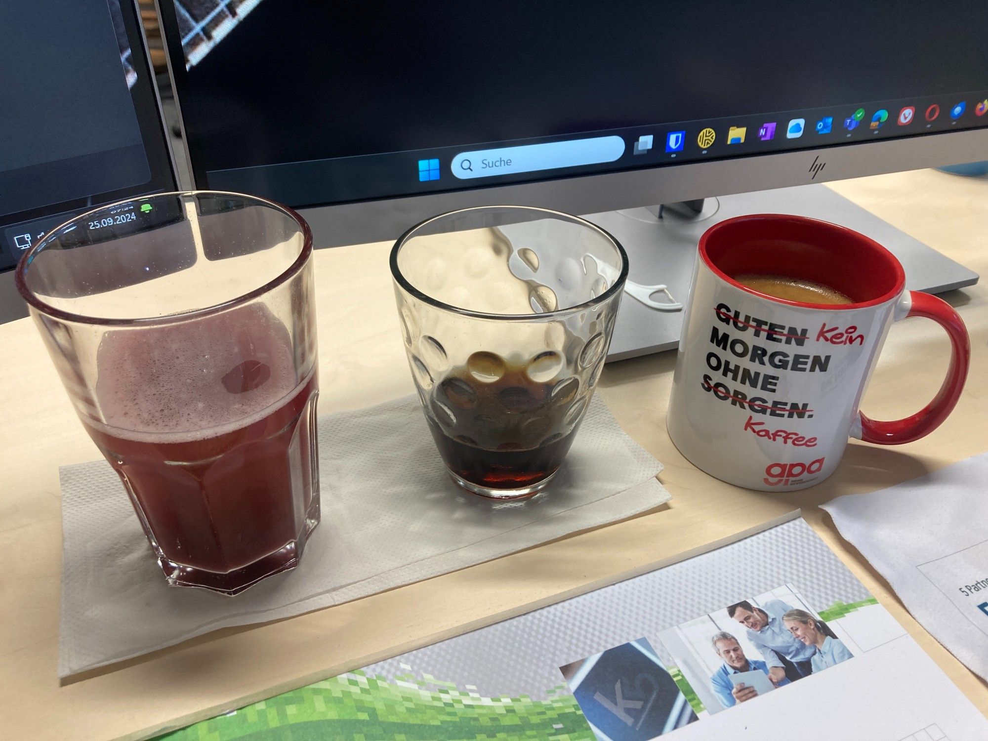 Auf dem Schreibtisch vor den Monitoren stehen ein größeres Glas mit dunkelroter Limo, ein fast leeres, kleineres Glas mit Cola und eine rot-weiße Tasse voller Kaffee.