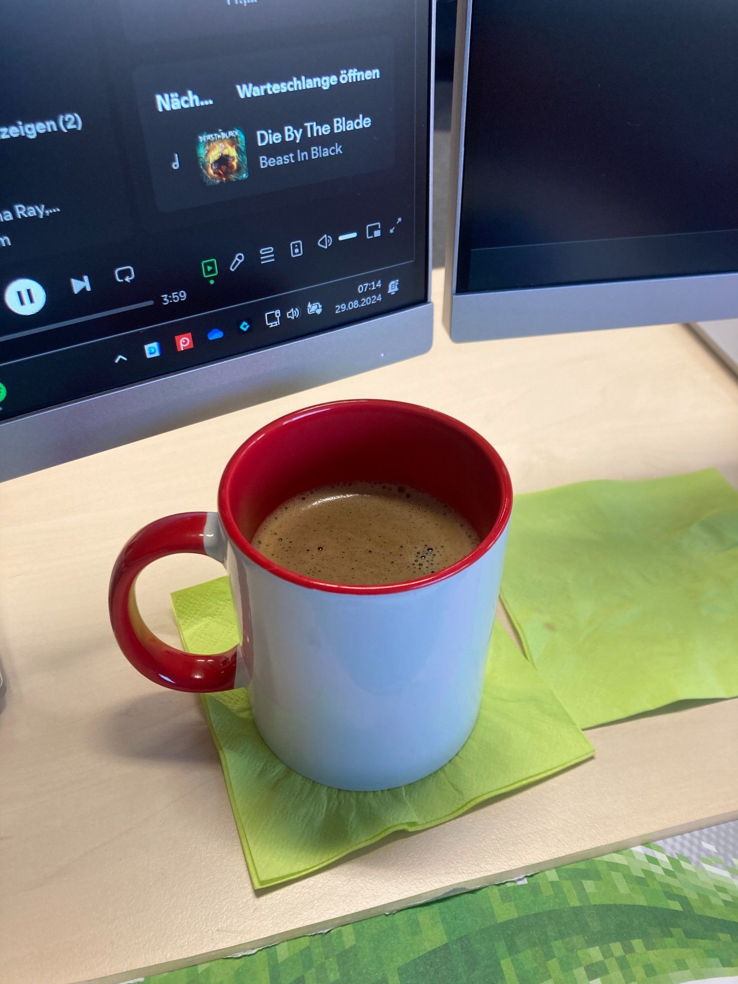 Große, weiße  Tasse mit roten Akzenten voller Kaffee auf dem Schreibtisch vor den Monitoren stehend.