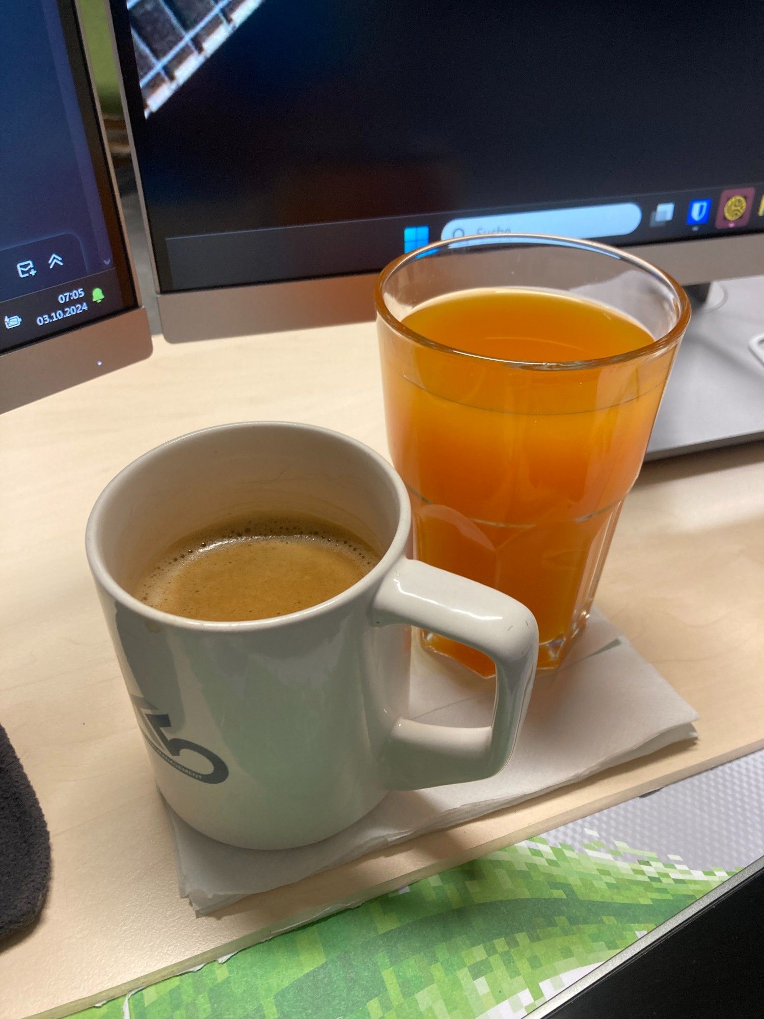 Große, weiße Tasse mit Kaffee und großes transparentes Glas mit orangener Limo am Schreibtisch vor den Monitoren stehend.