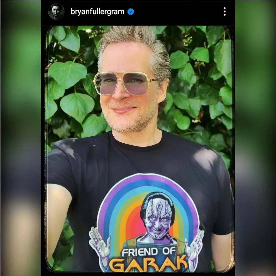 Bryan Fuller (who was a writer for DS9 and Voyager, and co-creator of Discovery) wearing a Friend of Garak t-shirt and aviator sunglasses. The shirt is black and has a drawing of Garak making jazz hands in front of a rainbow.