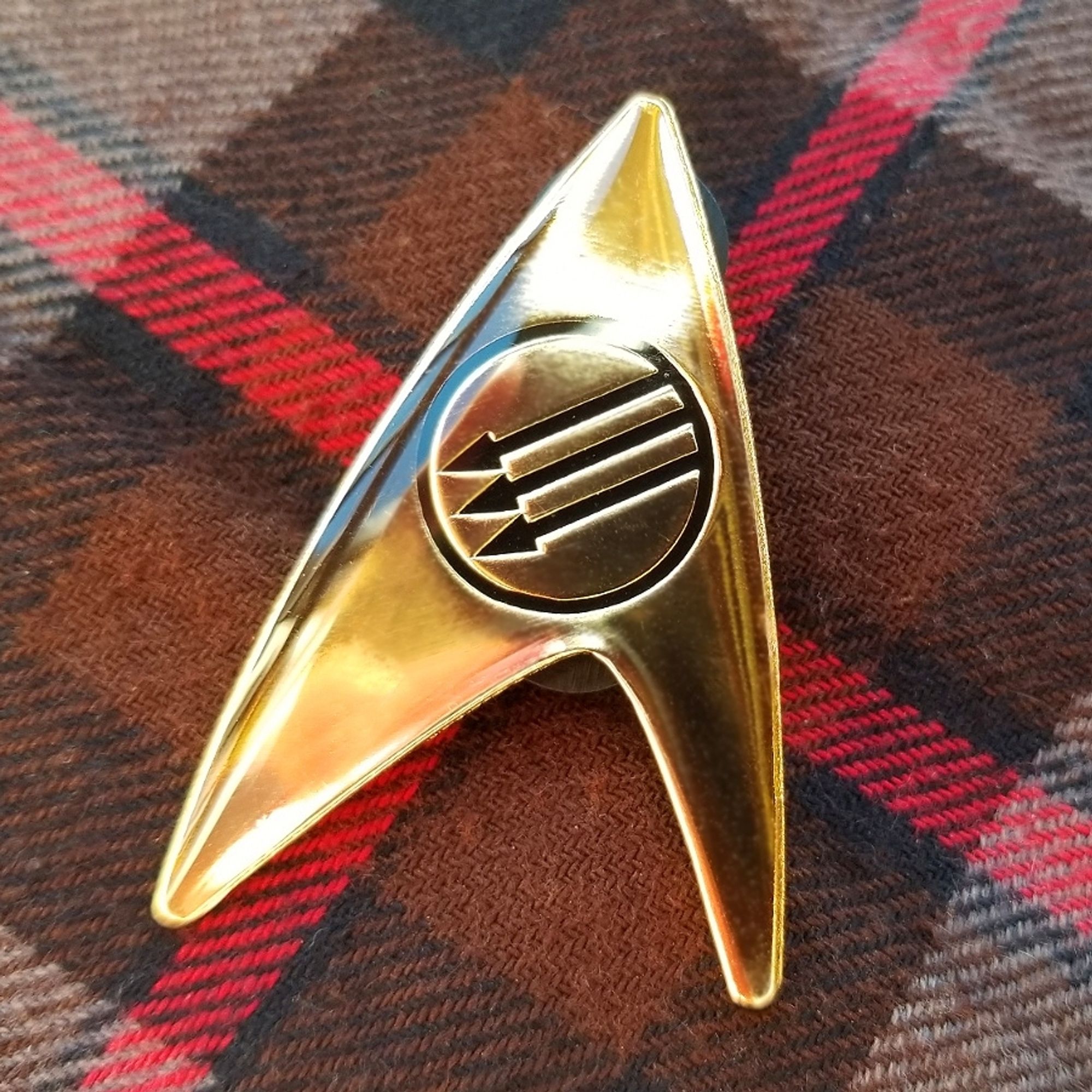 A Starfleet delta insignia pin in gold-colored metal and a black Three Arrows symbol in the center, and a high gloss finish.