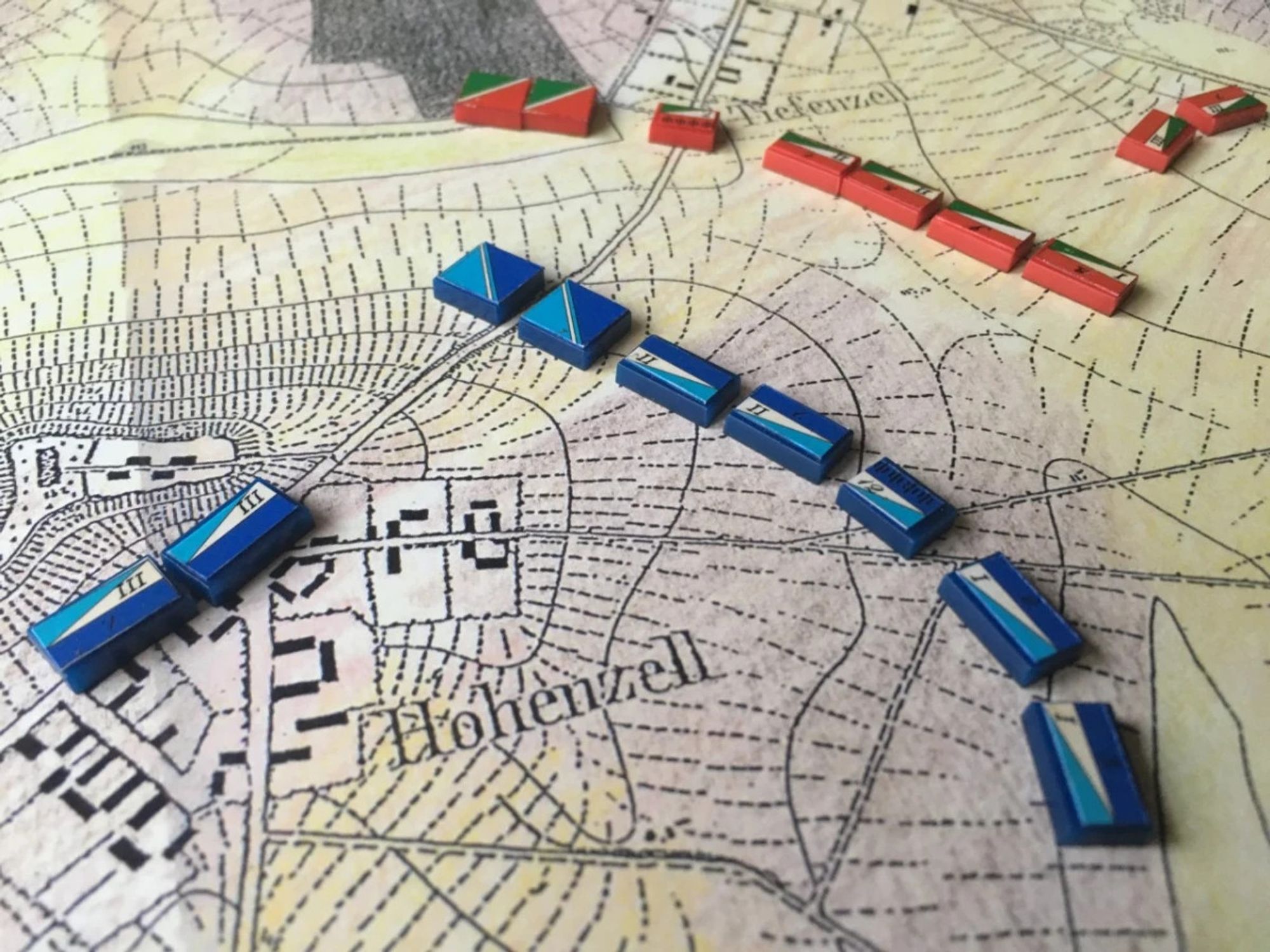 A close-up of a Kriegsspiel game. Tiny wooden rectangles painted in bright red and blue face off against each other in formation atop a printed map. The map depicts terrain features like roads, hills, and urban areas.