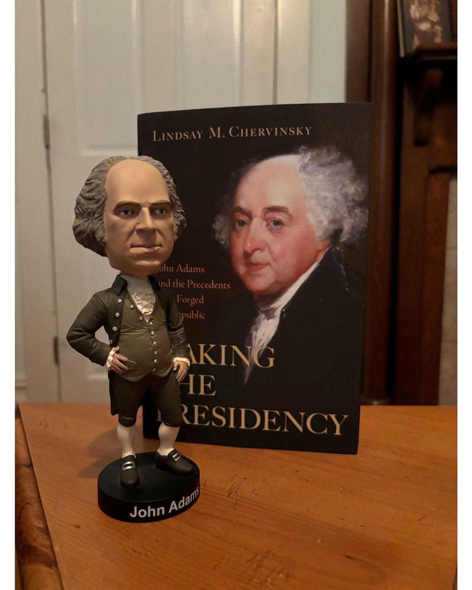 A John Adams bobblehead in front of a hardcover book on Adams’s presidency