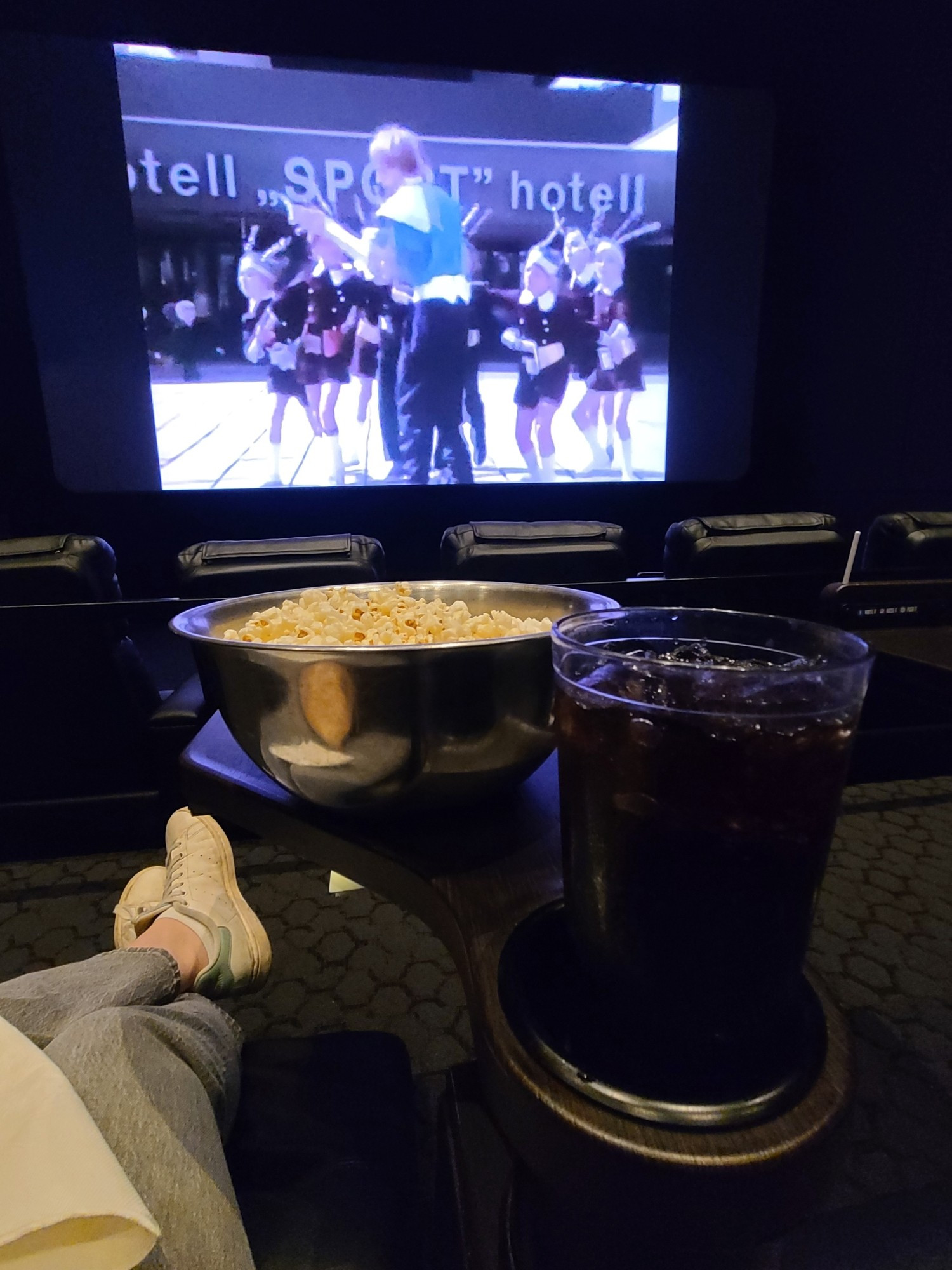 Movie theater screen playing some video involving robots, my legs in grey jeans and my feet in Stan Smiths, a metal mixing bowl of popcorn, a plastic tumbler of Dr. Pepper.