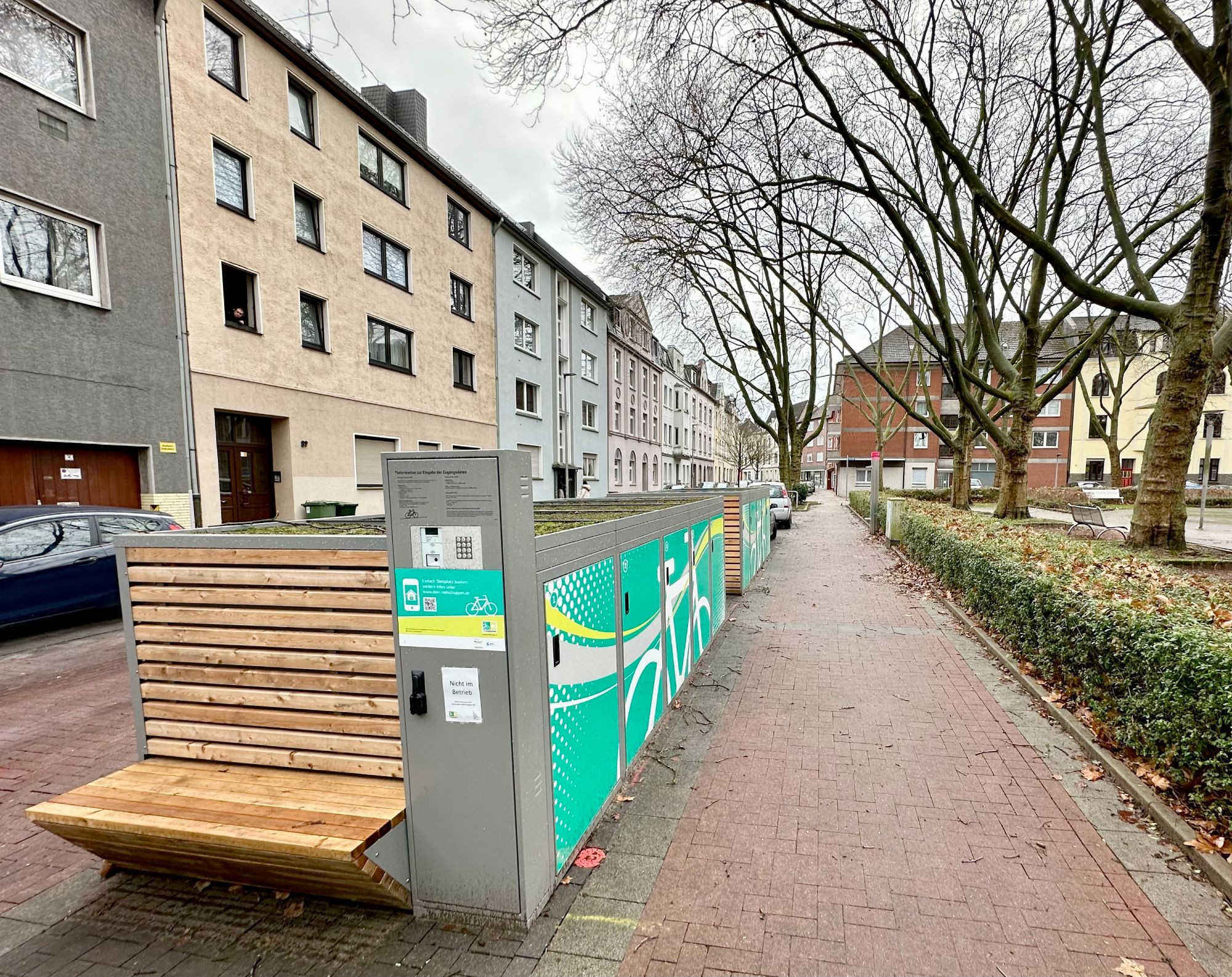 Links sind zwei Element von Fahrradboxen mit je 5 Stellplätzen zu sehen. Vor Kopf befindet sich ein Bedienterminal sowie eine Sitzbank aus Holz. Die Dächer sind begrünt. Insgesamt hat ein Element die Größe eines Autostellplatzes.