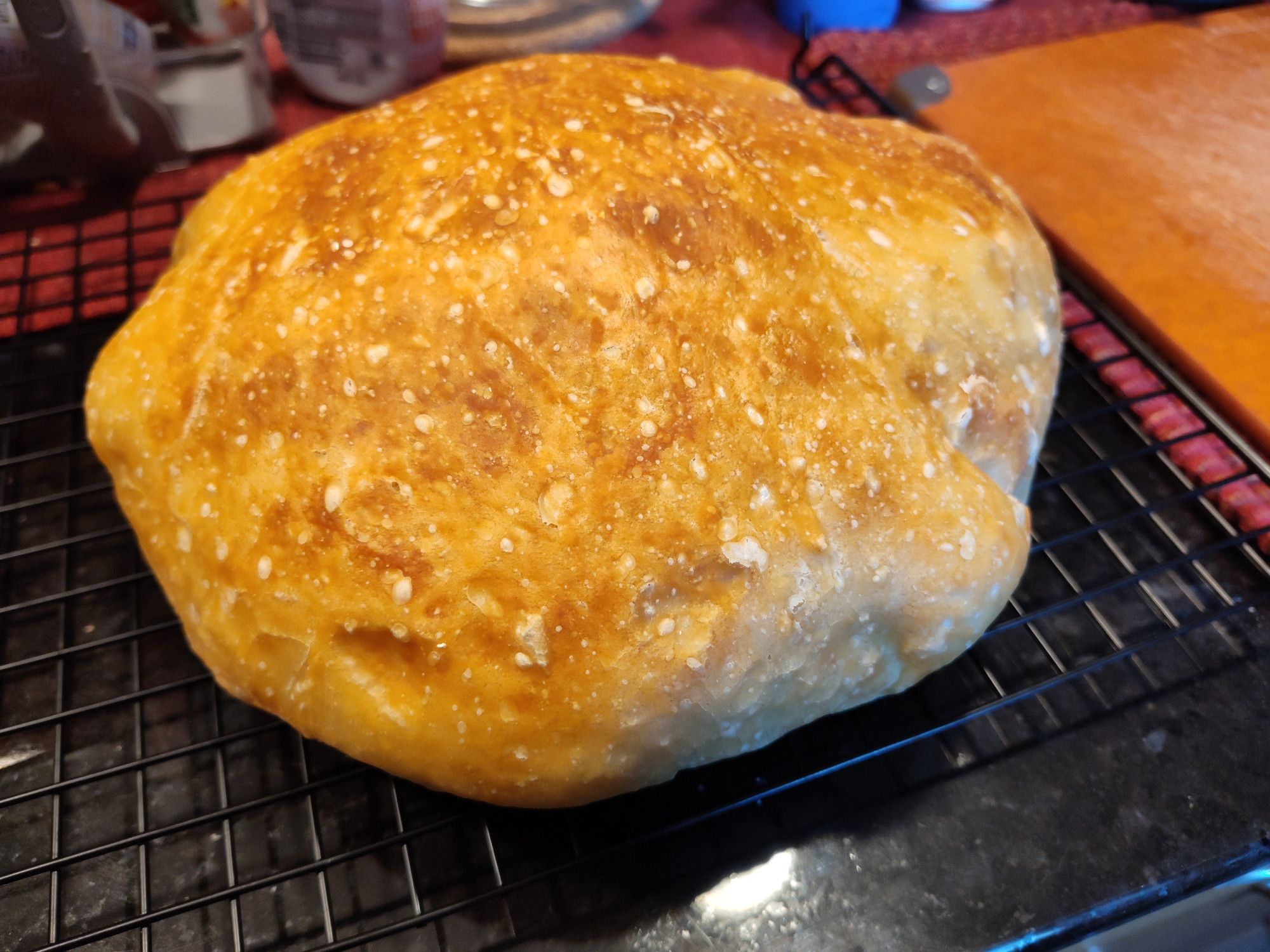 A circular loaf of white bread on a black cooling rack, the crust is a wonderful gold color, darkening to light brown here and there.