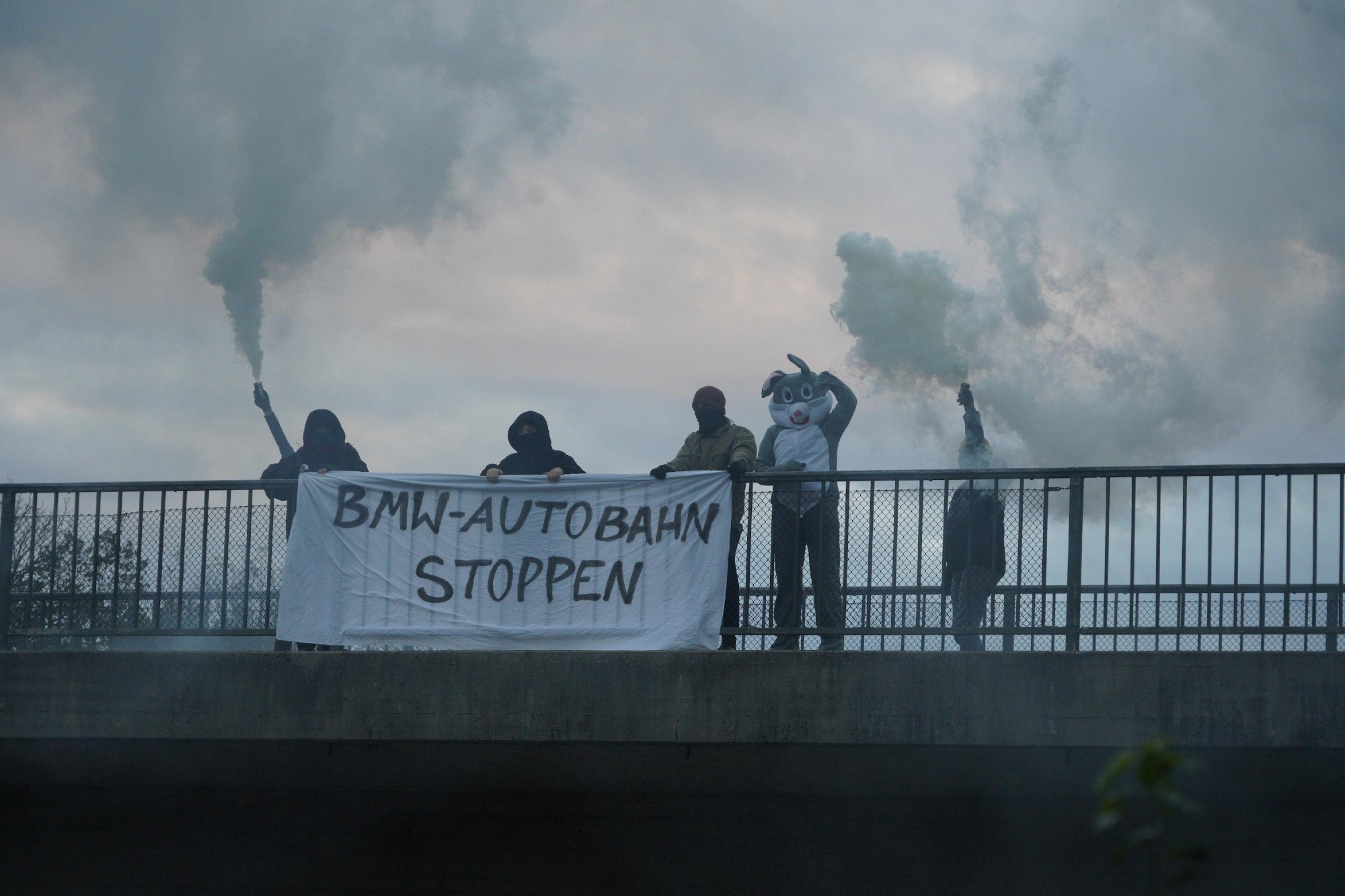 Menschen und ein Mensch mit Hasenkostüm halten auf einer Autobahnbrücke ein Banner "BMW-Autobahn stoppen" und halten Rauchtöpfe