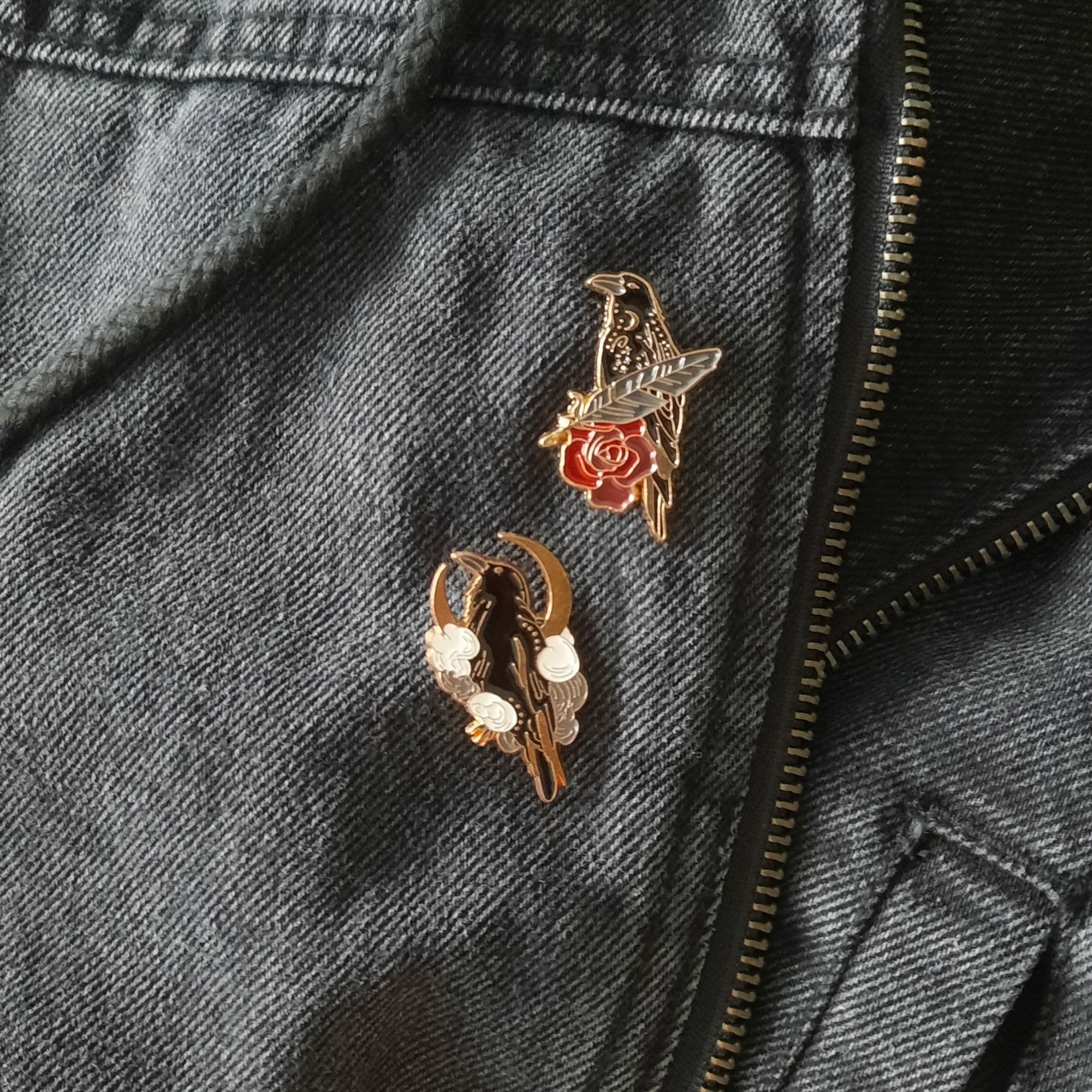 Photo of a part of my black jeans jacket, showing two enamel pins depicting crows, the line art is gold, one crow is depicted with a feather and a red rose while the other has a crescent moon behind it, sitting in the middle of smoke or clouds