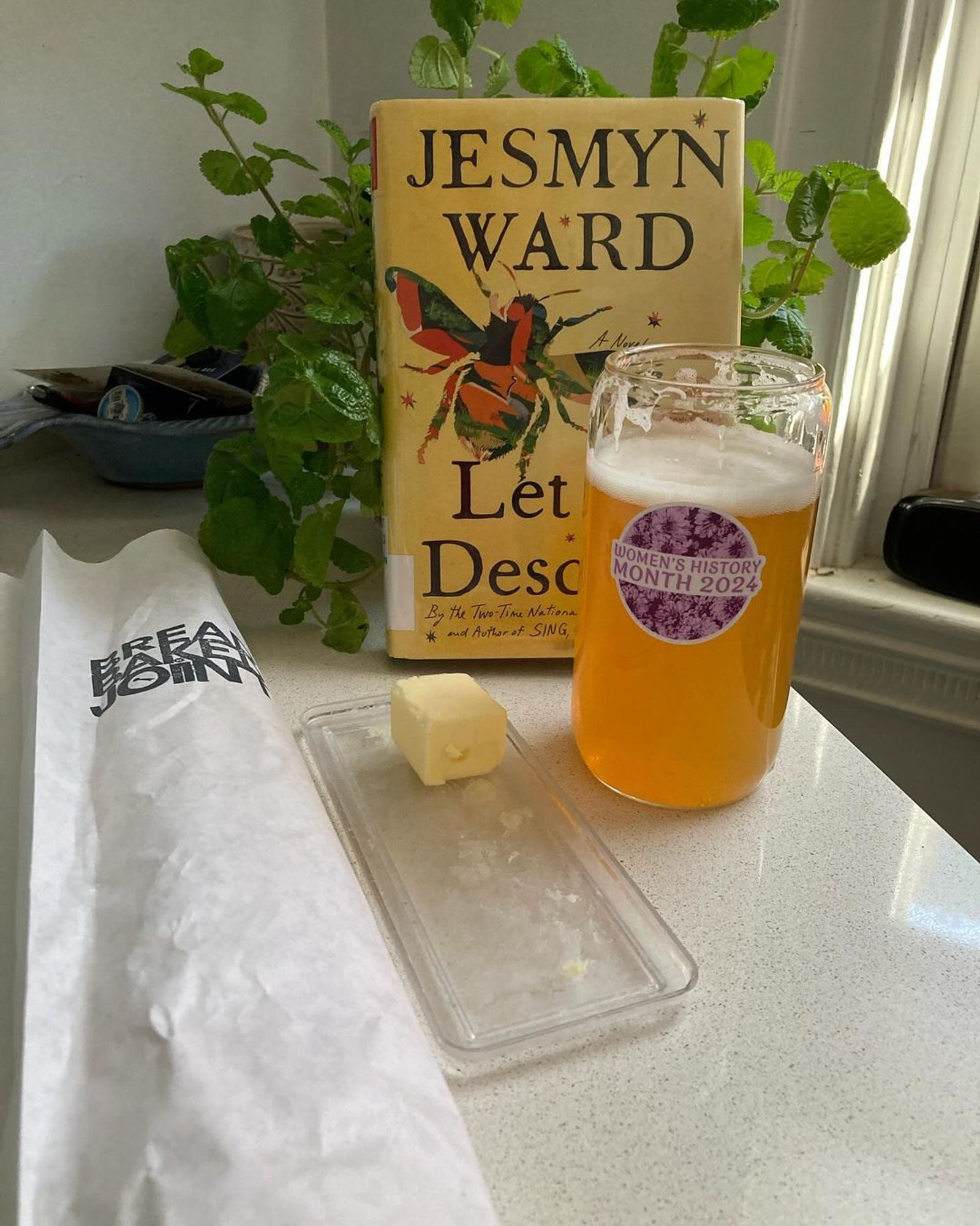 On a white counter rests a white bag (containing an excellent baguette) that reads, "Bread Baked Joint", a little bit of butter on a clear plastic plate, a clear class with a "Women's History Month 2024," Jesmyn Ward's *Let Us Descend" upright, and a green plant.