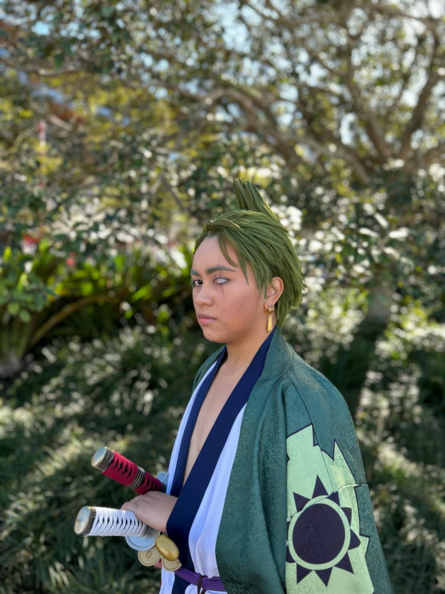 Photo of a person cosplaying as Zoro from One Piece in his Wano arc outfit. He is posed with a hand on one of his swords, staring at the camera with a serious expression, in a sunny outdoor setting.