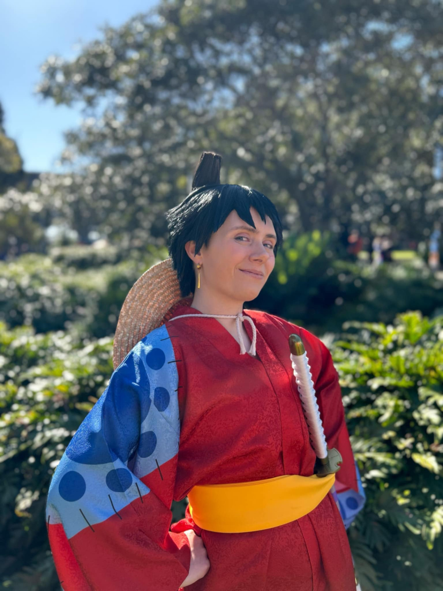 Photo of a person cosplaying as Luffy from One Piece in his Wano arc outfit. They are posed with their hands on their hips, looking at the camera with a confident smile, in a sunny outdoor setting.