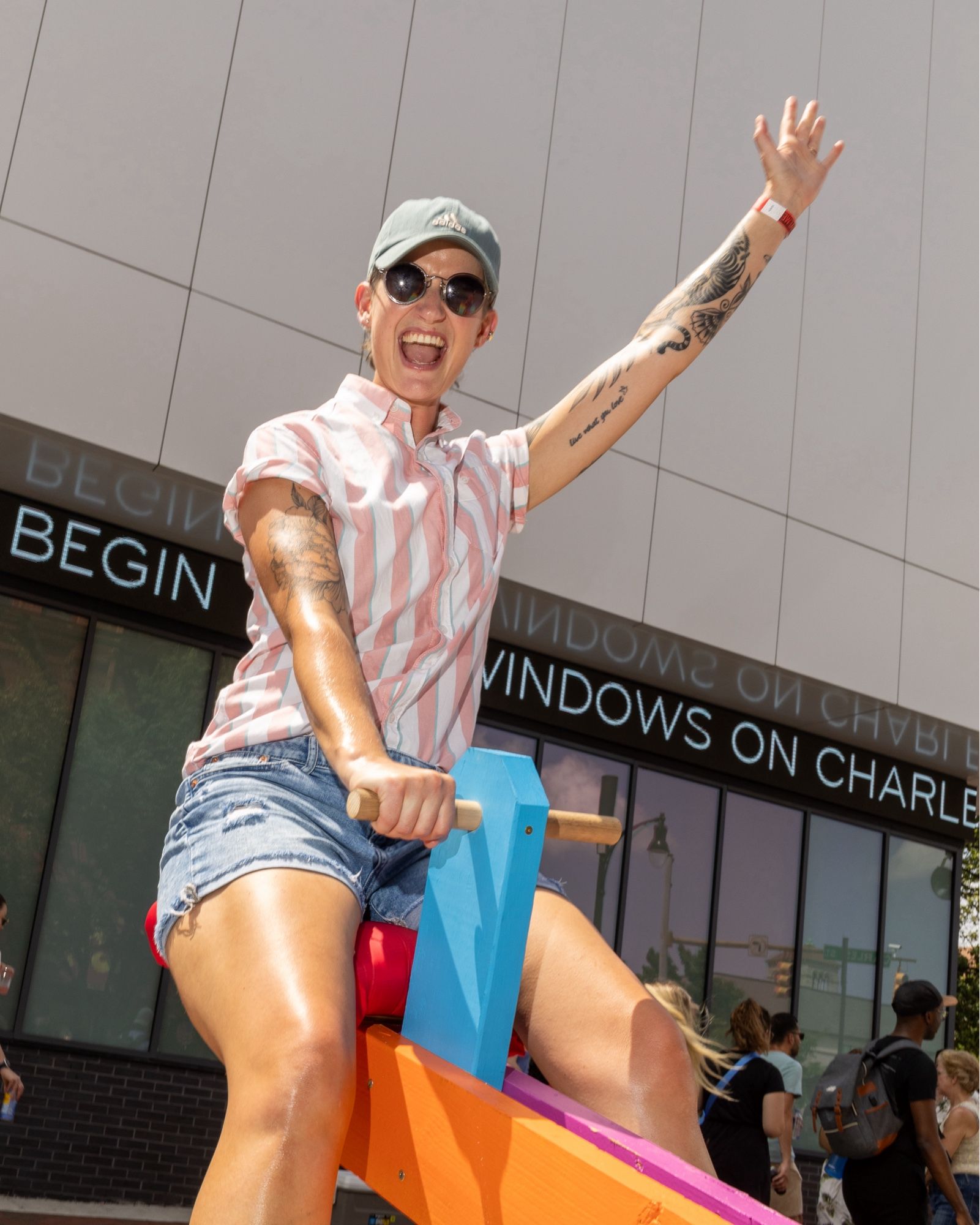 color photo of a person on one end of a multi-colored see saw. They are up at the maximum height the see saw will go and they are holding one arm way up above their head and have a huge smile on their face. They are wearing sunglasses, a pink ad white short sleeved shirt, and cut off denim shorts.