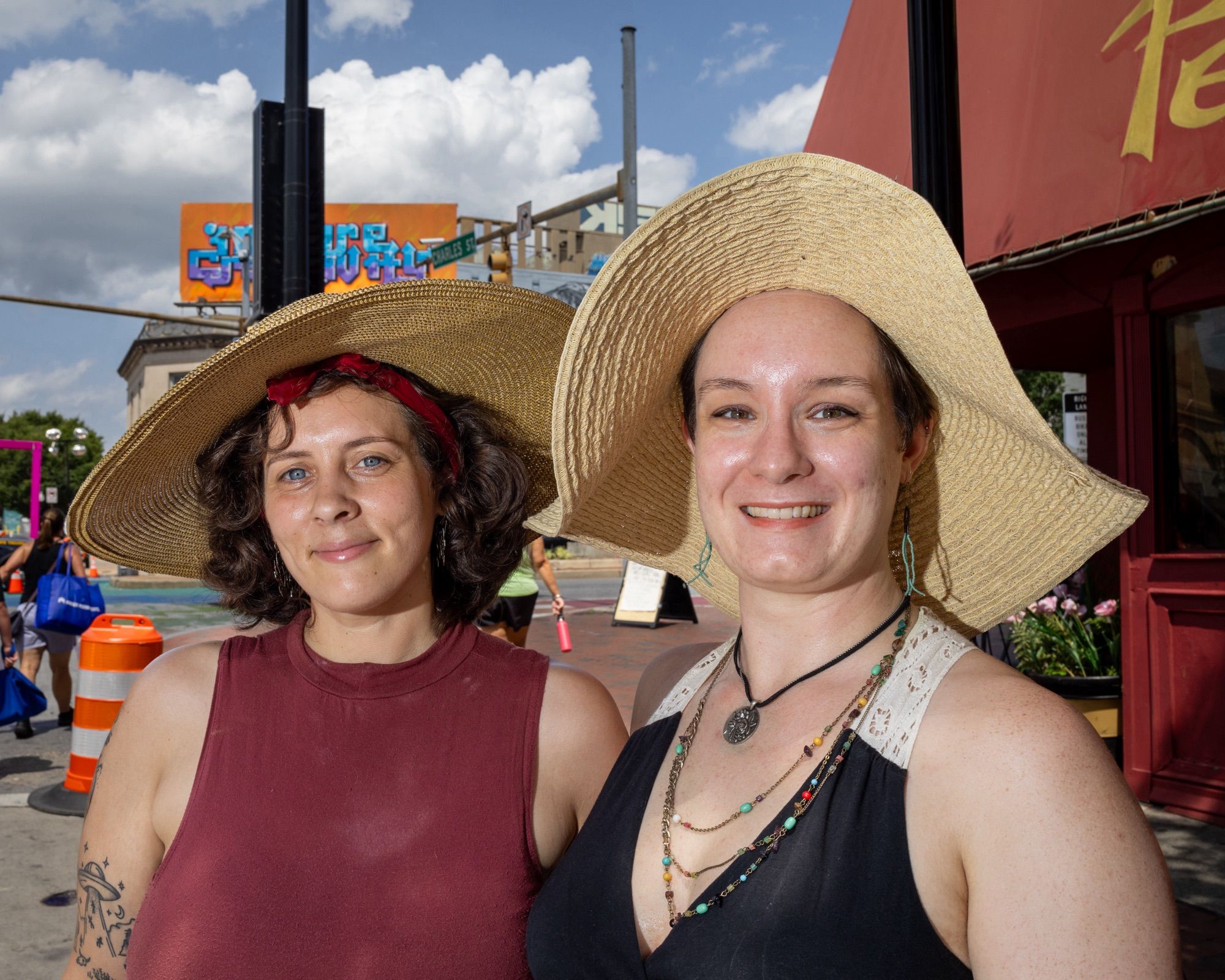 Color photo of two people wearing large floppy straw hats. They are both wearing sleeveless tops. THey are both smiling at the camera.