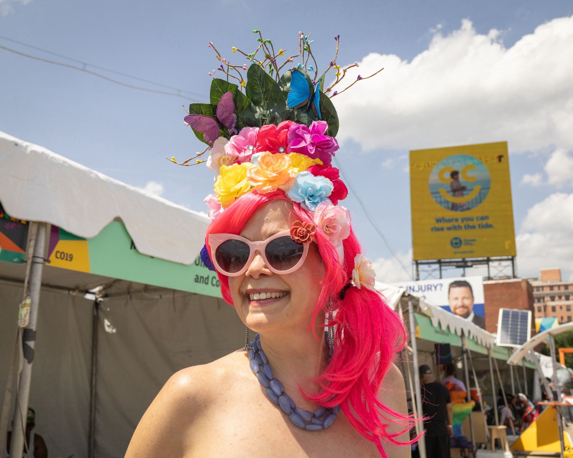 Color photo of a person with long magenta hair (a wig, maybe?) and a tall flower arrangement for a headpiece. They are smiling and wearing pink framed sunglasses and a semi-chunky blue necklace. There are event tents behind them and a sunny sky with fluffy white clouds in the background.