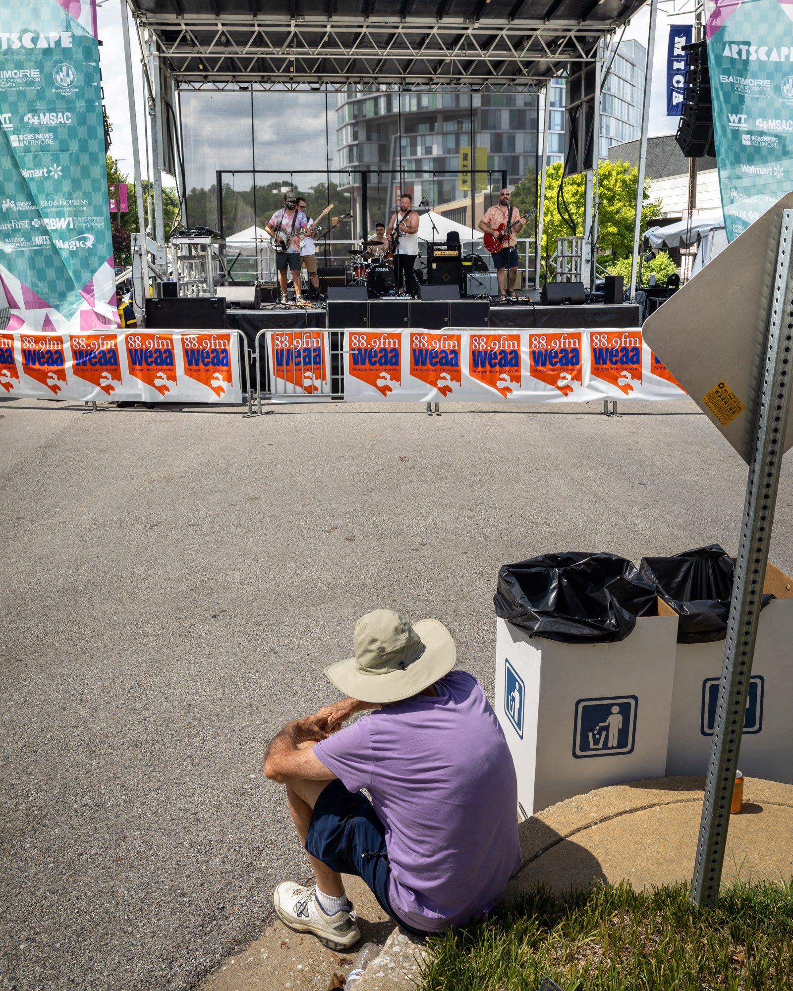 color photo of an outdoor stage with five piece rock band performing a song. There is crowd control fencing in front of the stage but there is only one person visible in the photo that is looking at and listening to the band. The solitary audience member is well back from the stage, sitting on a curved  with their arms wrapped around their knees. The space between them and the band is empty pavement.