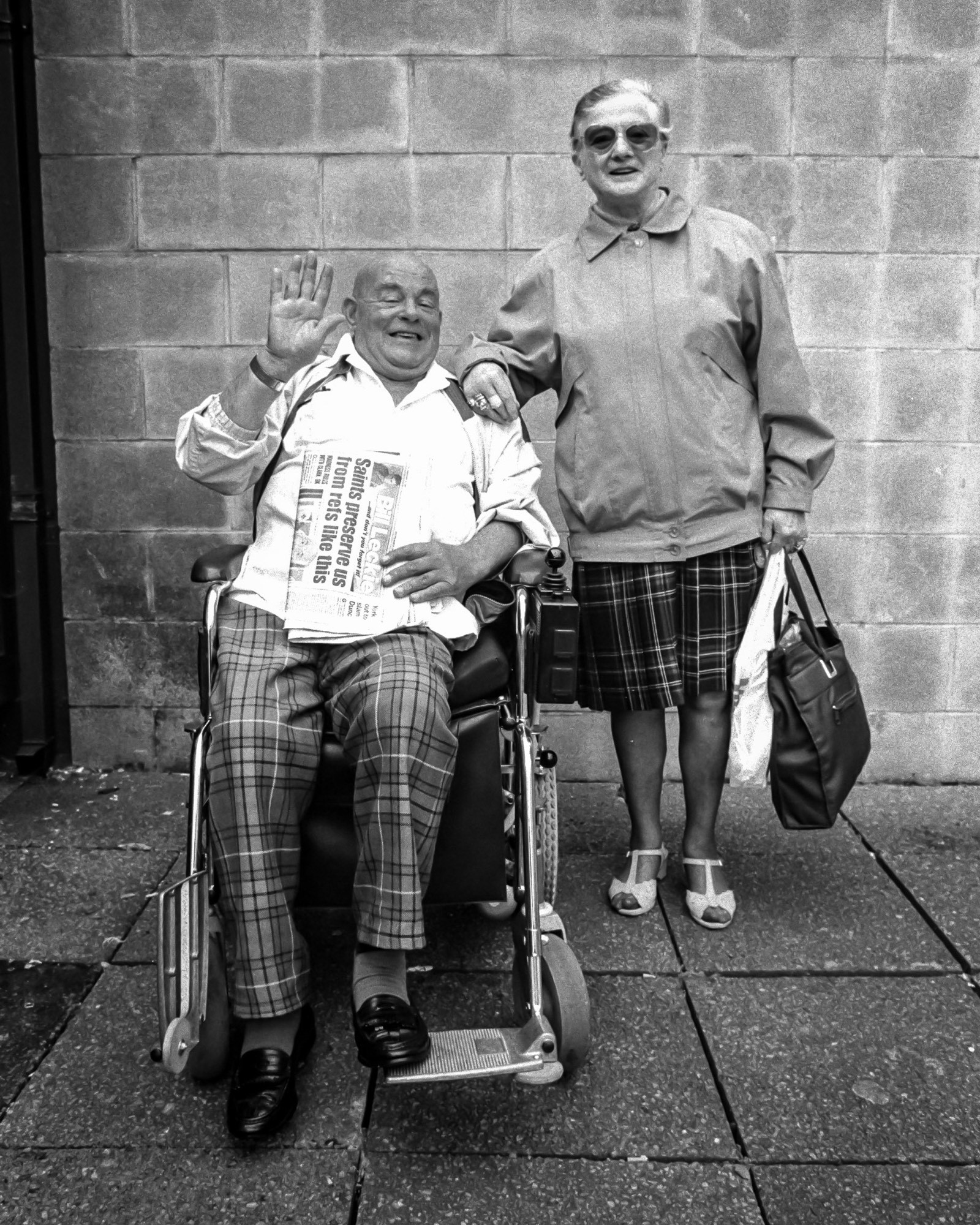 Black and white photo of an elderly man a woman full length facing the camera on a sidewalk. The man is in a wheelchair with a newspaper on his lap. He is bald, smiling, and has his hand up waving at the photographer. The woman to his side is also smiling. She is standing with one hand on the man’s shoulder and from the other hangs a purse and a shopping bag. He is wearing plaid pants and she is wearing a plaid skirt. Behind them is a cinder block wall.