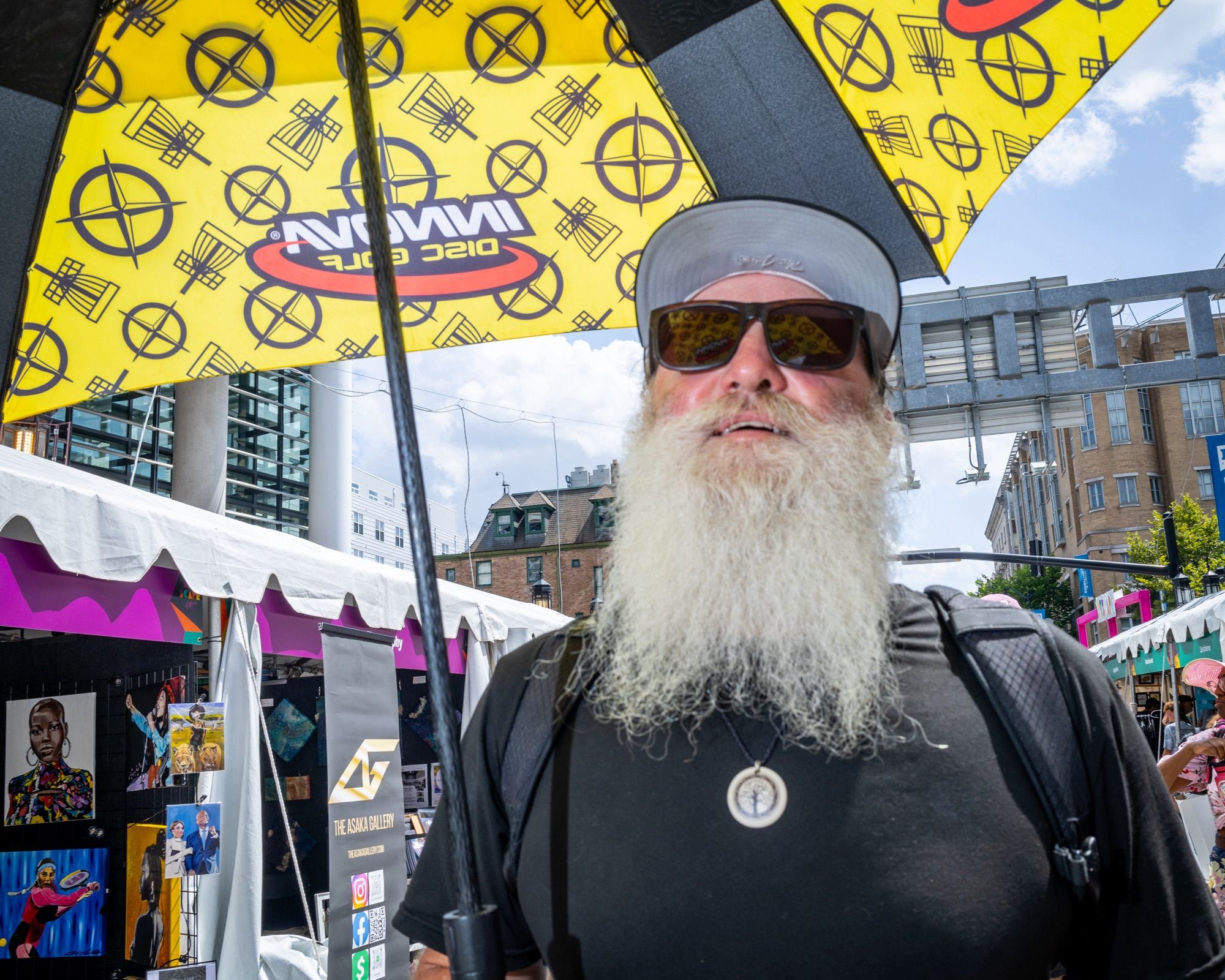 Head and shoulder portrait of a person in a black t-shirt holding a yellow umbrella (they are using it as a parasol) that is partly cropped out of the photo. They have a long white beard and are wearing sunglasses and a cap. They are smiling. Behind are street fair tents and some apartment buildings.