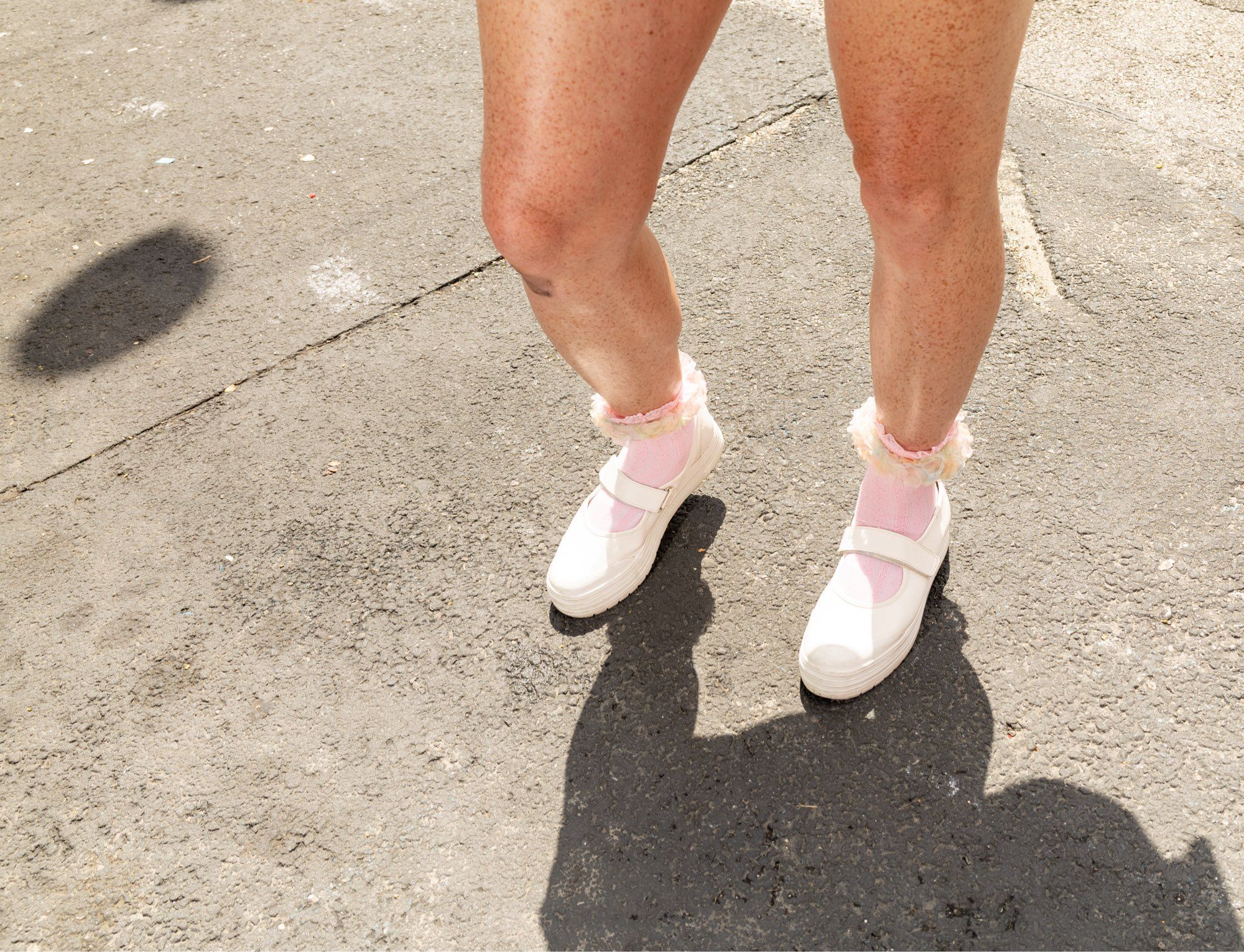 Bennie’s backlit, shiny, freckled legs. They are wearing white Mary Jane sneakers and light pink socks with ruffles on top. Their shadow is cast forward and out of the frame. To the left, a single round shadow of a balloon hovers on the sidewalk.