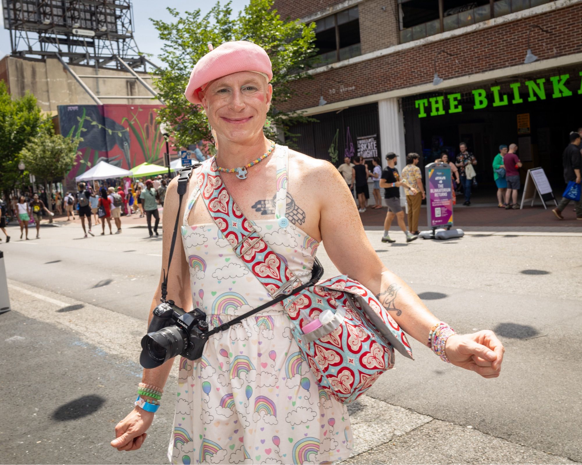 Street portrait of Benjie, who is smiling at the camera. They are wearing a white sleeveless dress with a rainbow print pattern. The framing is a ¾ view. They have a digital camera and a shoulder bag (with a heart pattern) slung over one shoulder. They are wearing a pink beret. They have a necklace made of pastel colored stars and a single blue charm of a hobby horse in the middle. Behind them, other street fair goers are passing by with some city buildings beyond.