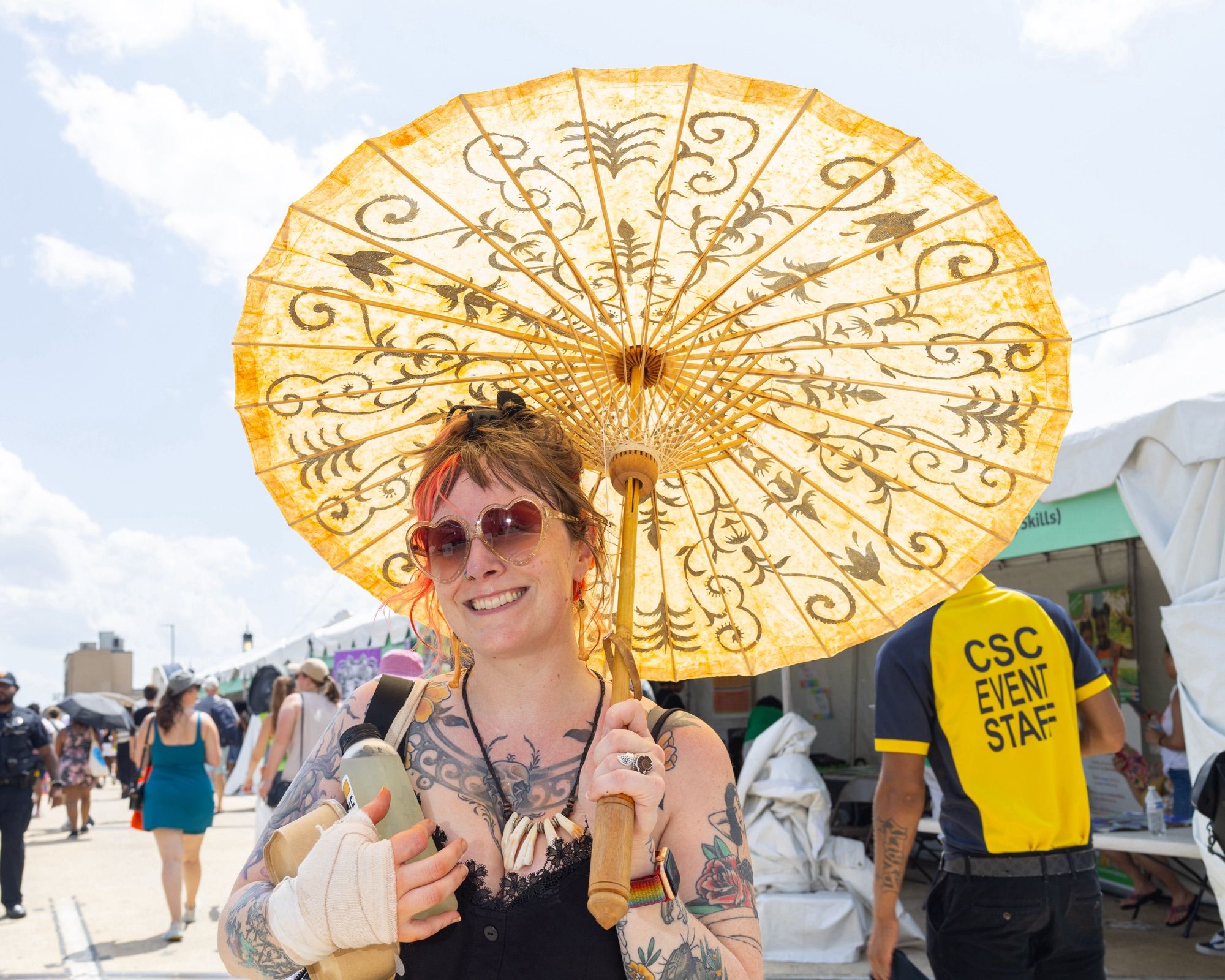 Color photo of a person holding a yellow parasol on a sunny day at a street fair. They are smiling and wearing heart-shaped sunglasses. They are also wearing a black spaghetti strap dress (visible from the chest up) and have a bandage on their right hand. Behind them are a number of people walking and a bright, partly cloudy sky.